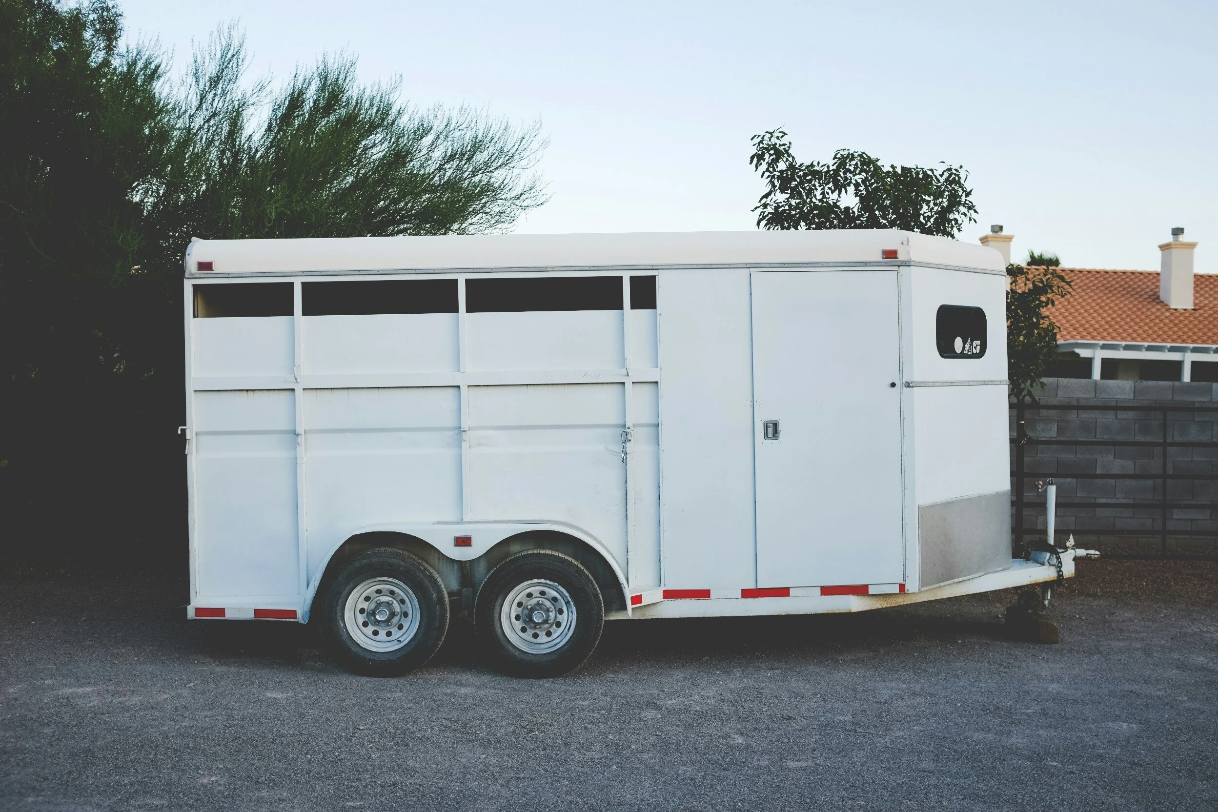 White horse trailer parked on a gravel driveway, surrounded by trees and a brick wall.