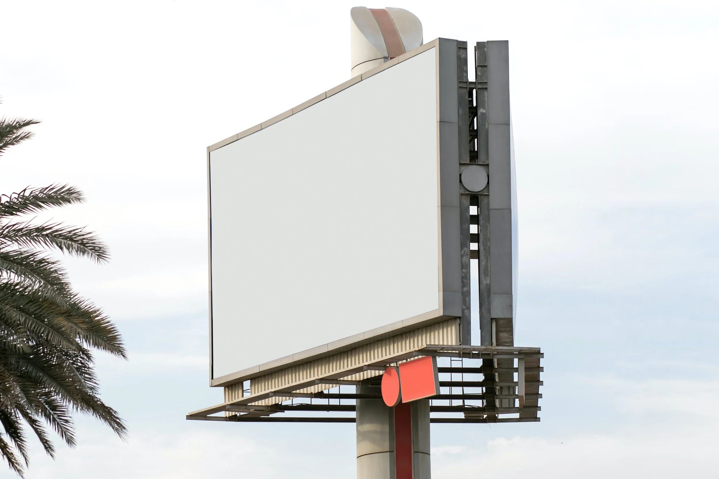 Blank billboard with a metal frame next to a palm tree against a cloudy sky.