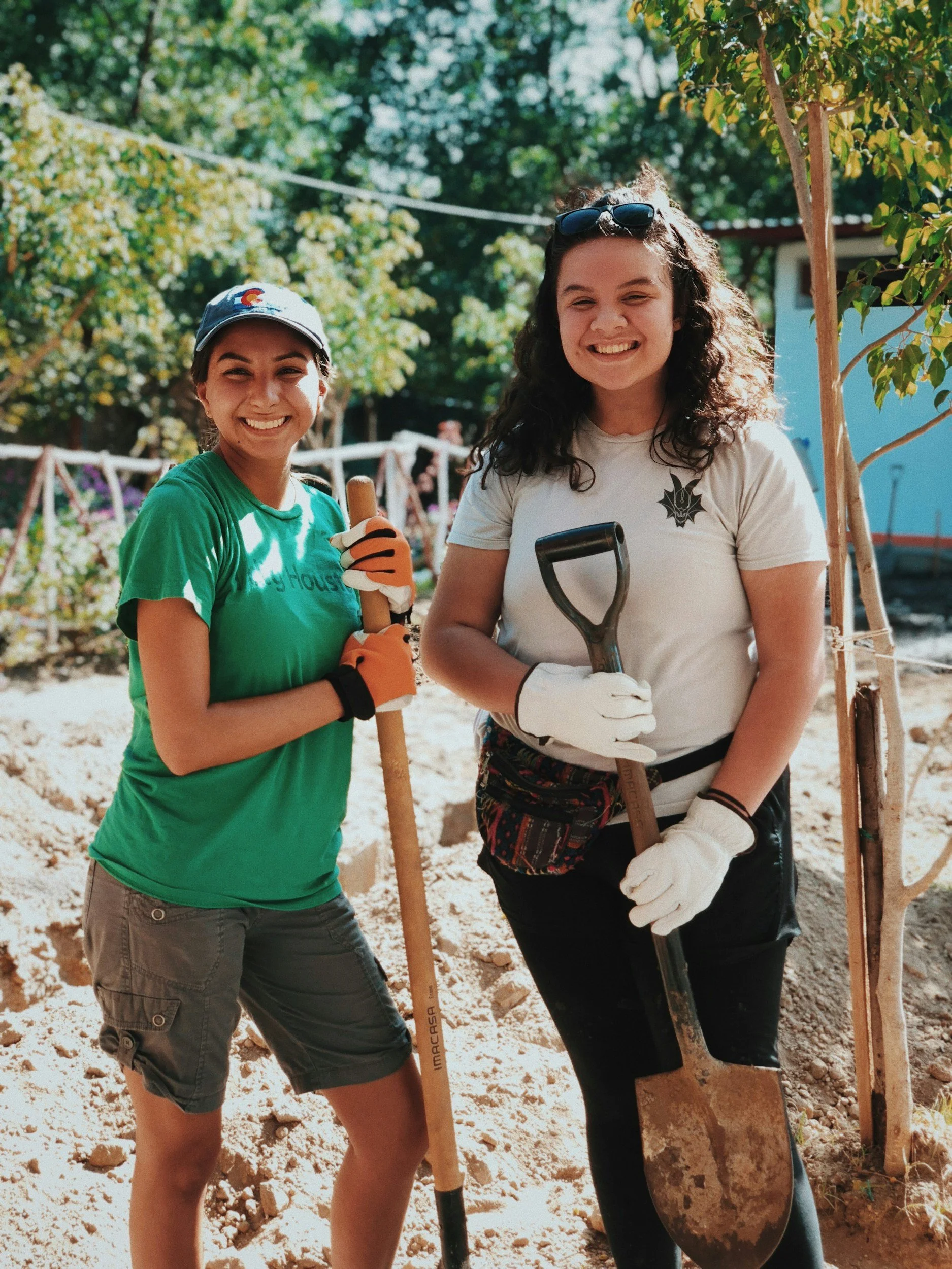Two girls smile at the camera holding a shovel and rake outside.