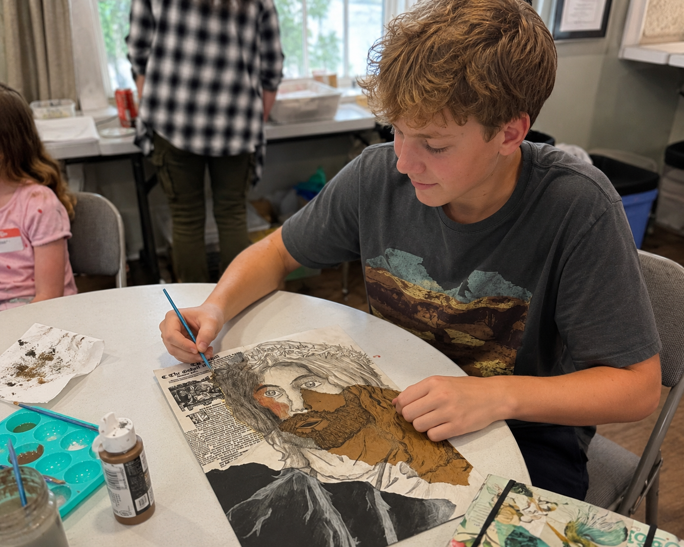 A student sits at a table working on an art peace with a pen in hand and a smile on his face.