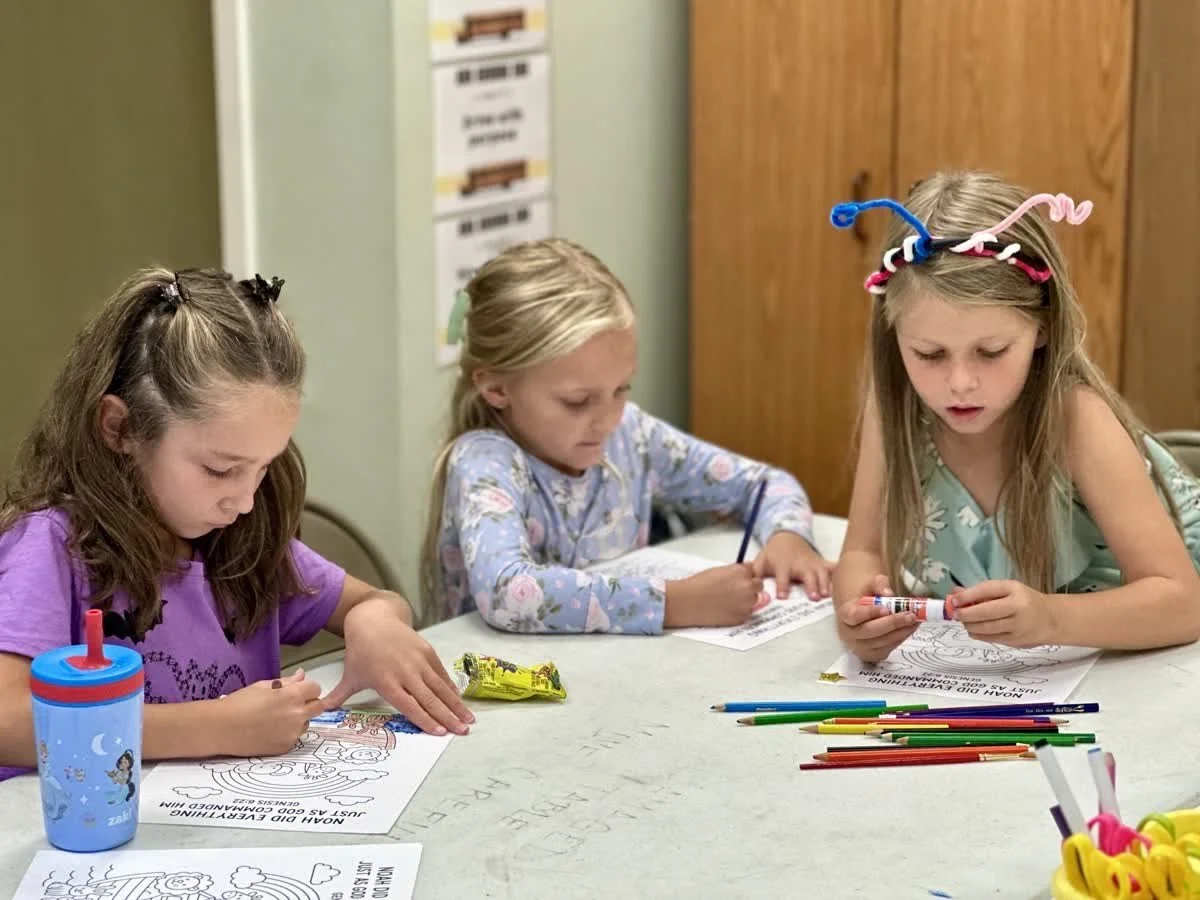 Three girls sit at a table, coloring on coloring sheets together with colored pencils.