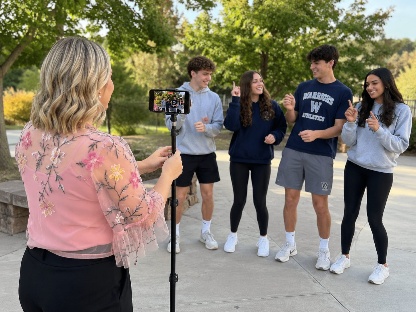 Teacher holds a a selfie stick with phone video on, while students do a social media reel in front of it.