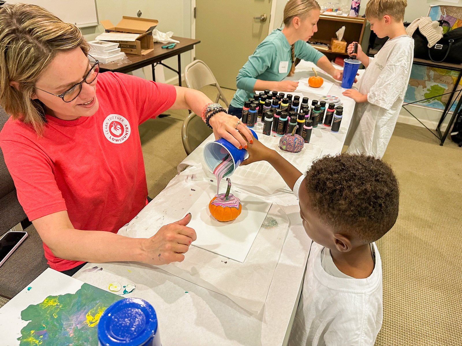 A teacher helps a student pour paint onto a pumpkin in art class.
