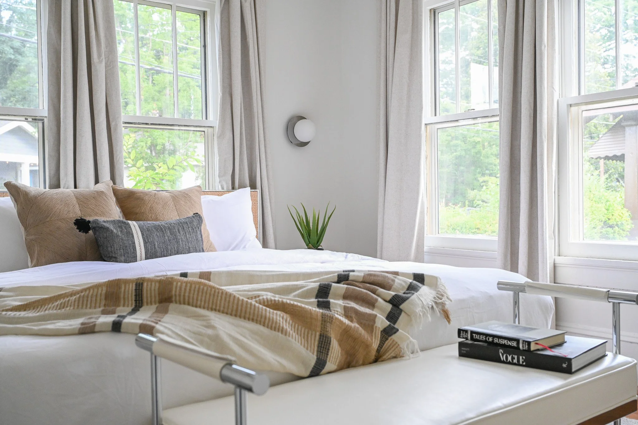 Bright bedroom with large windows, beige bedding, and a stack of books on a white bench at the foot of the bed.