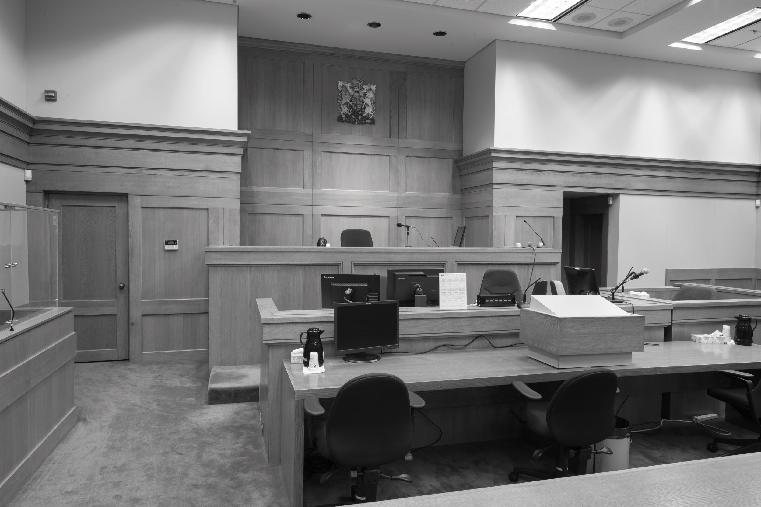 A courtroom with wooden paneling on the walls, a judge's bench at the front with microphones and chairs, and multiple computer monitors on the tables. The room is empty with no people present.