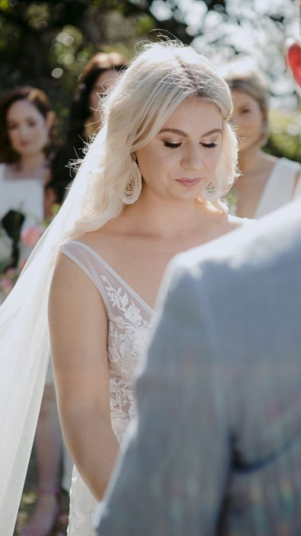 A bride and groom standing outdoors during their wedding ceremony, with bridesmaids in the background, trees, and sunlight.