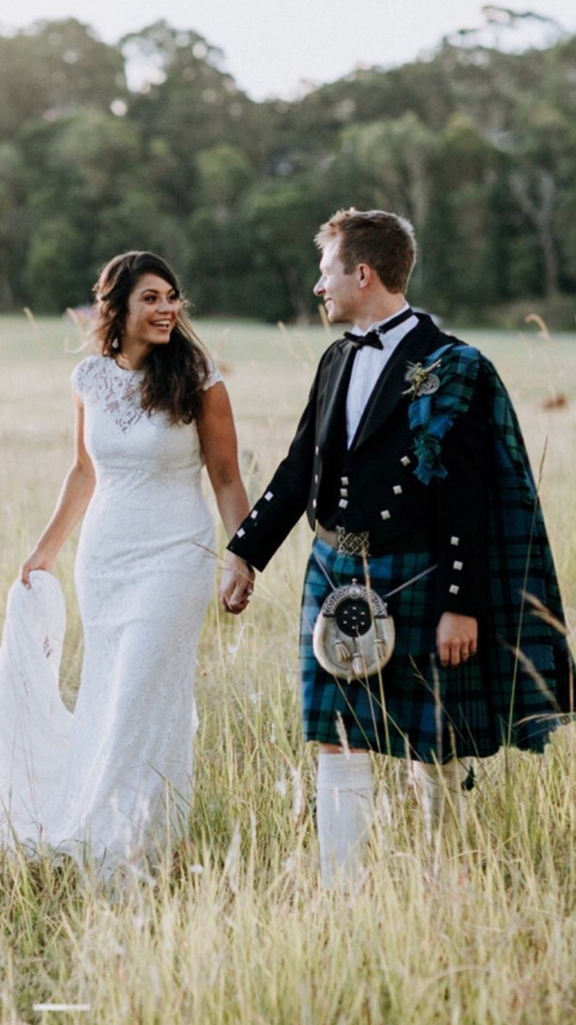 A bride and groom holding hands in a field, smiling at each other, with trees and hills in the background.