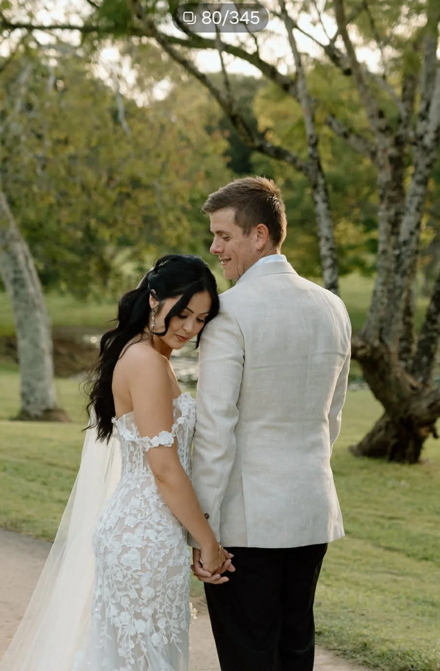 A bride in a white wedding gown holding a bouquet, standing outdoors on a grassy field with seating and a white trailer in the background, under a partly cloudy sky with sunlight.