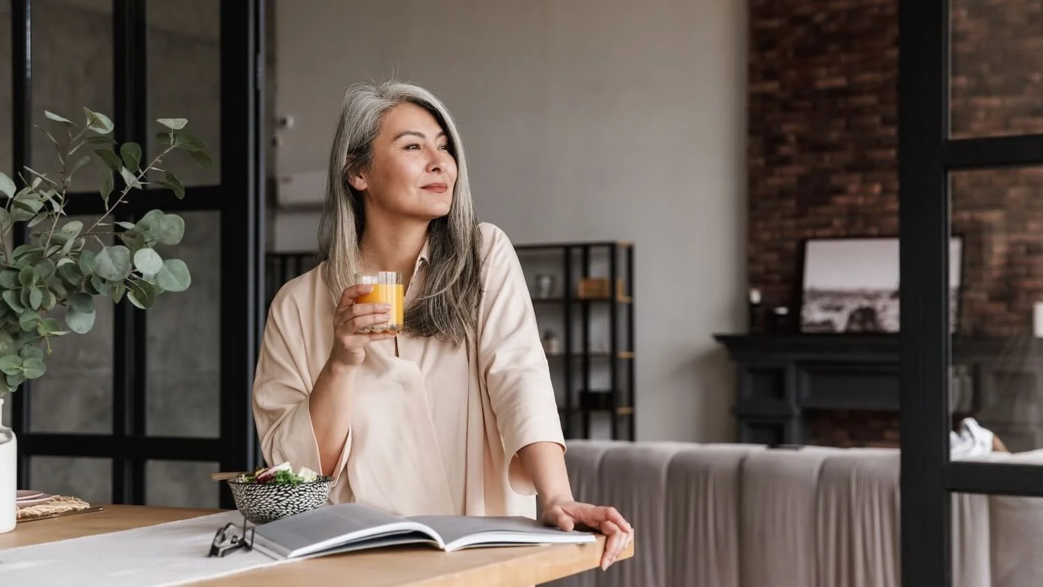 Turning 60 (Woman over 60 gazing outside as she drinks orange juice standing at counter)