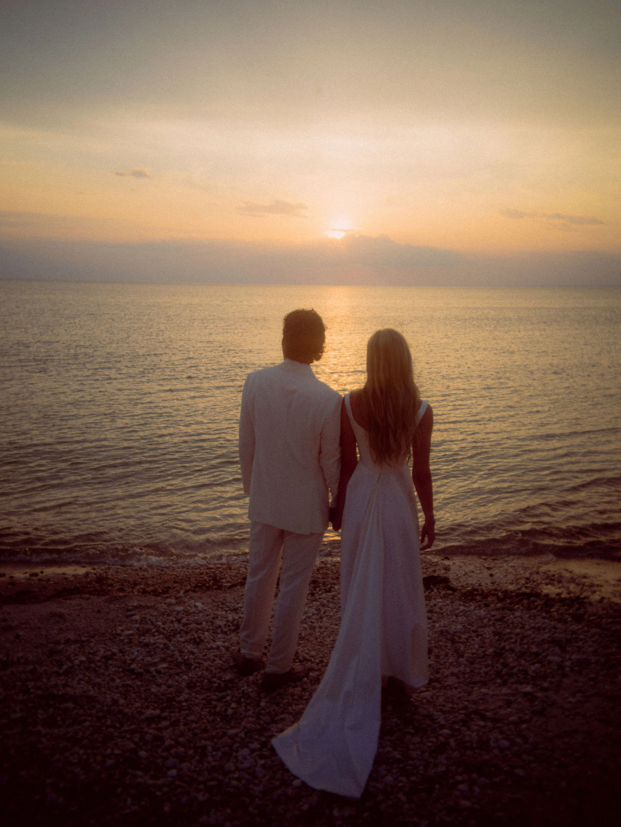 A couple holding hands and facing the ocean at sunset, with the woman wearing a white dress and the man in a light-colored suit.