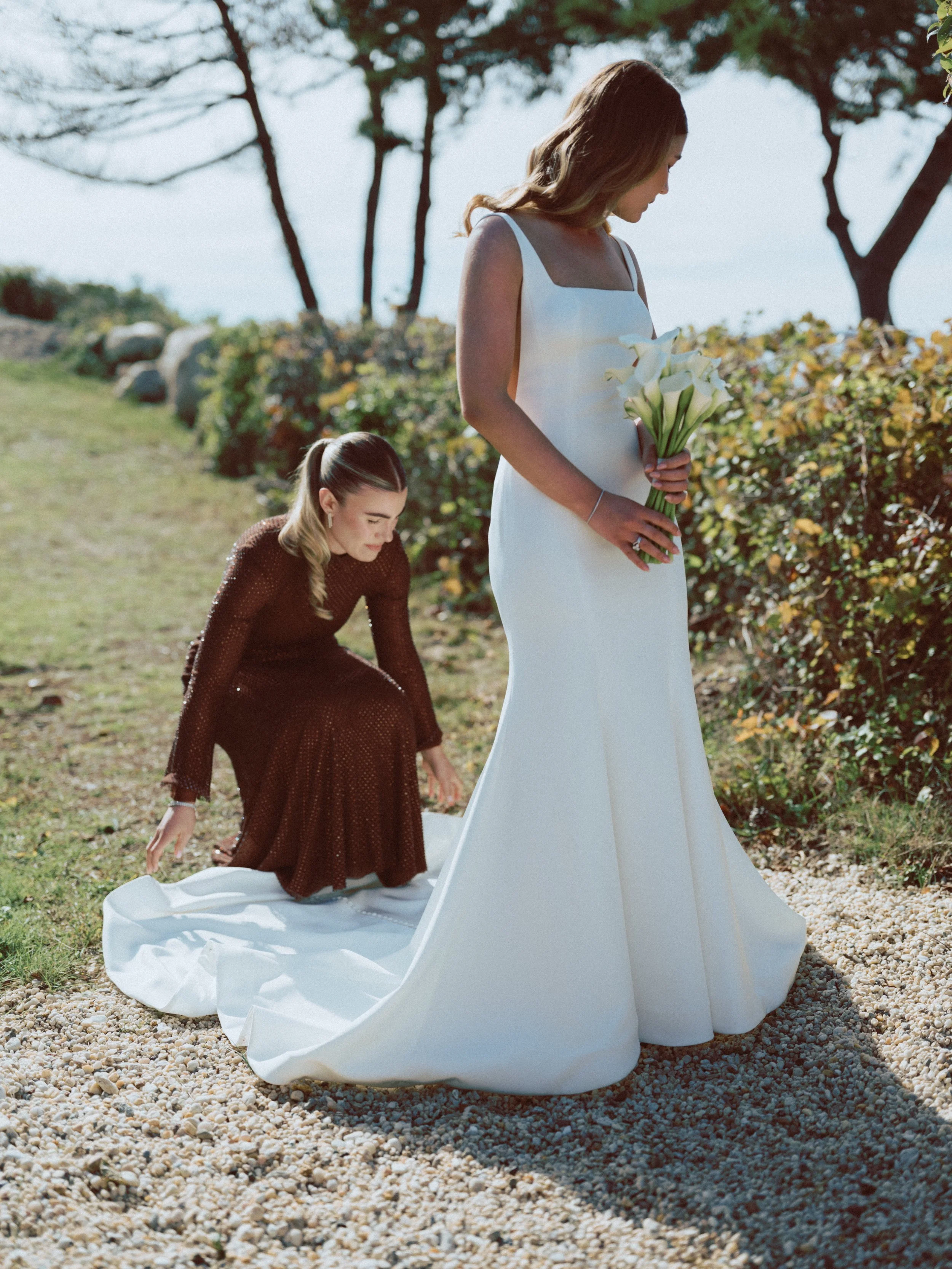 A bride in a white wedding dress holding a bouquet of white calla lilies, while another woman in a brown dress adjusts the train of her gown outdoors on a sunny day near trees and bushes.