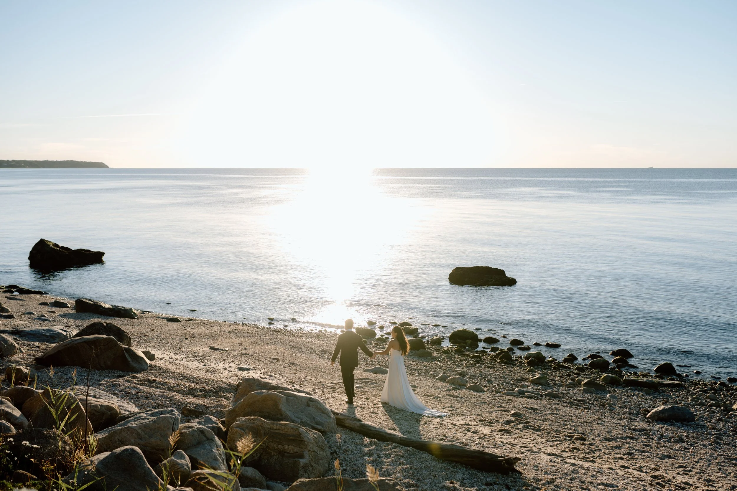 A bride and groom walking hand in hand on a beach at sunset, with rocks and calm water in the background.