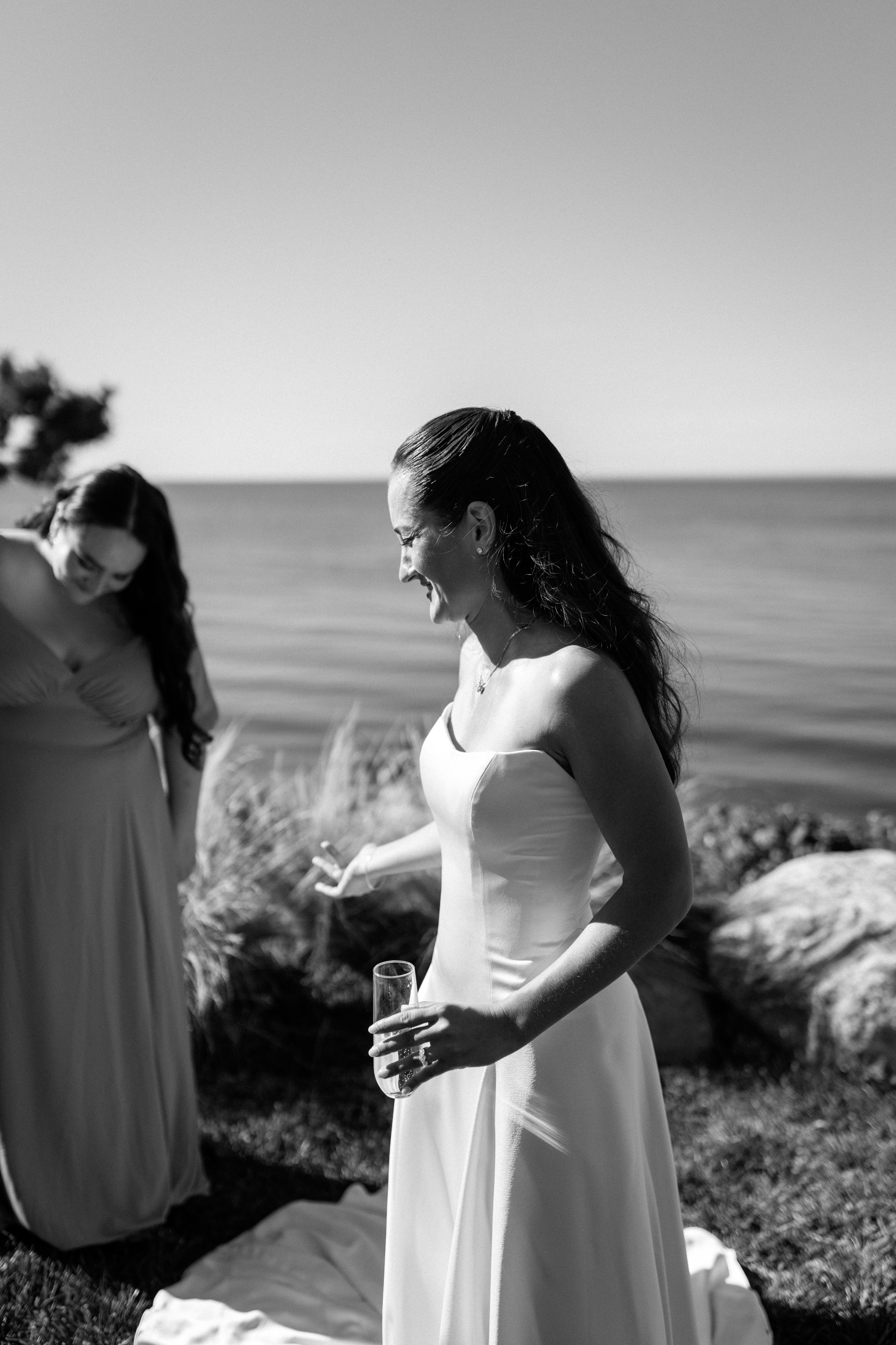 A woman in a wedding dress holding a glass and smiling outdoors near water, with another woman in a dress in the background.