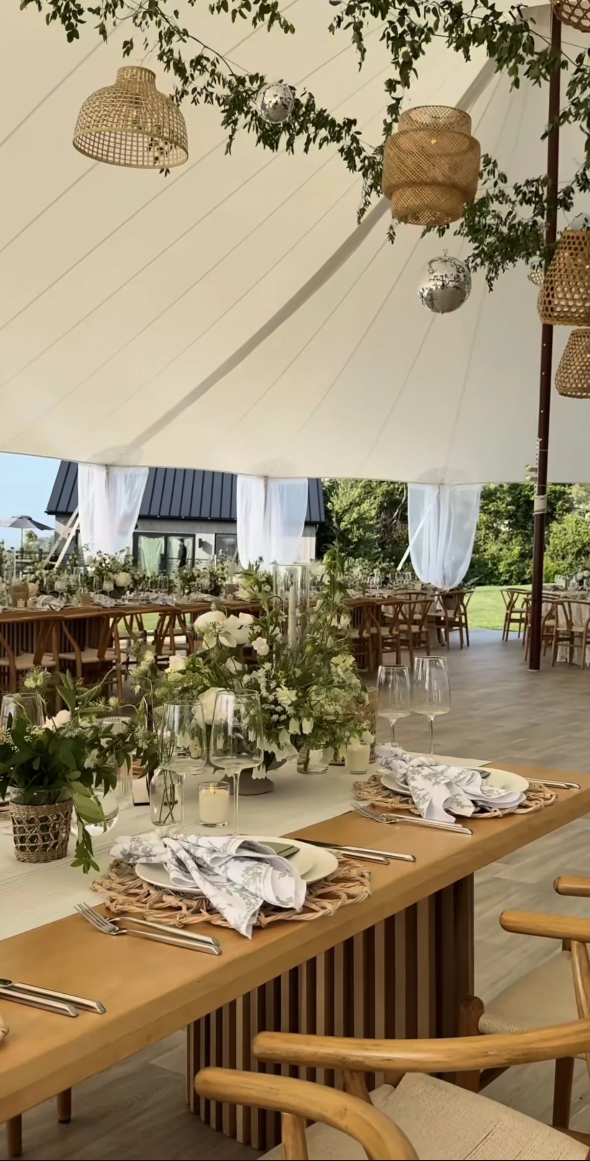 Indoor event space decorated for a gathering with long wooden tables, floral centerpieces, and neatly arranged table settings with plates, napkins, and glassware, under a large white canopy with hanging wicker and metal decorations.
