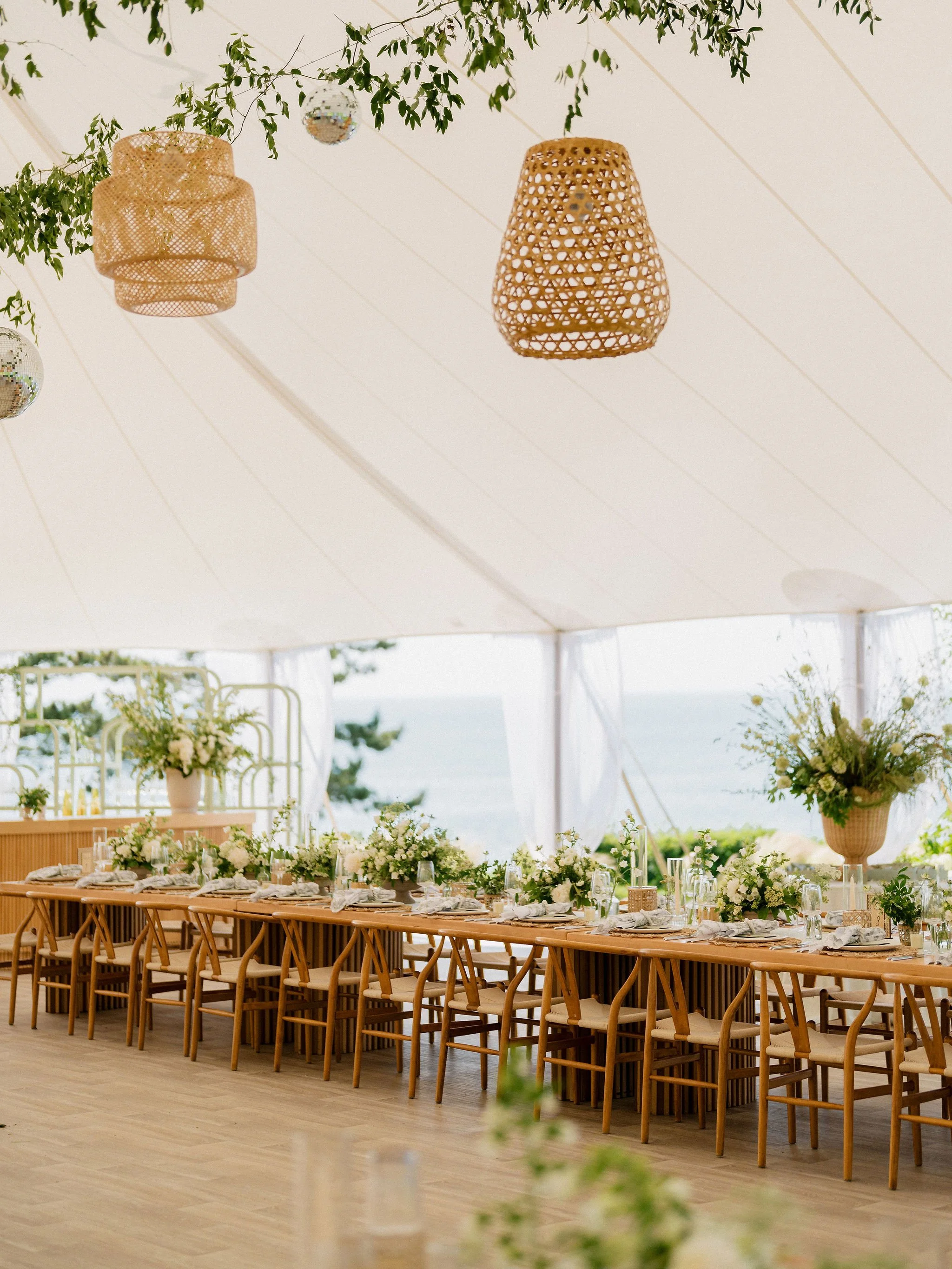 Decorated outdoor event tent with long wooden table, white floral arrangements, and hanging wicker lanterns.