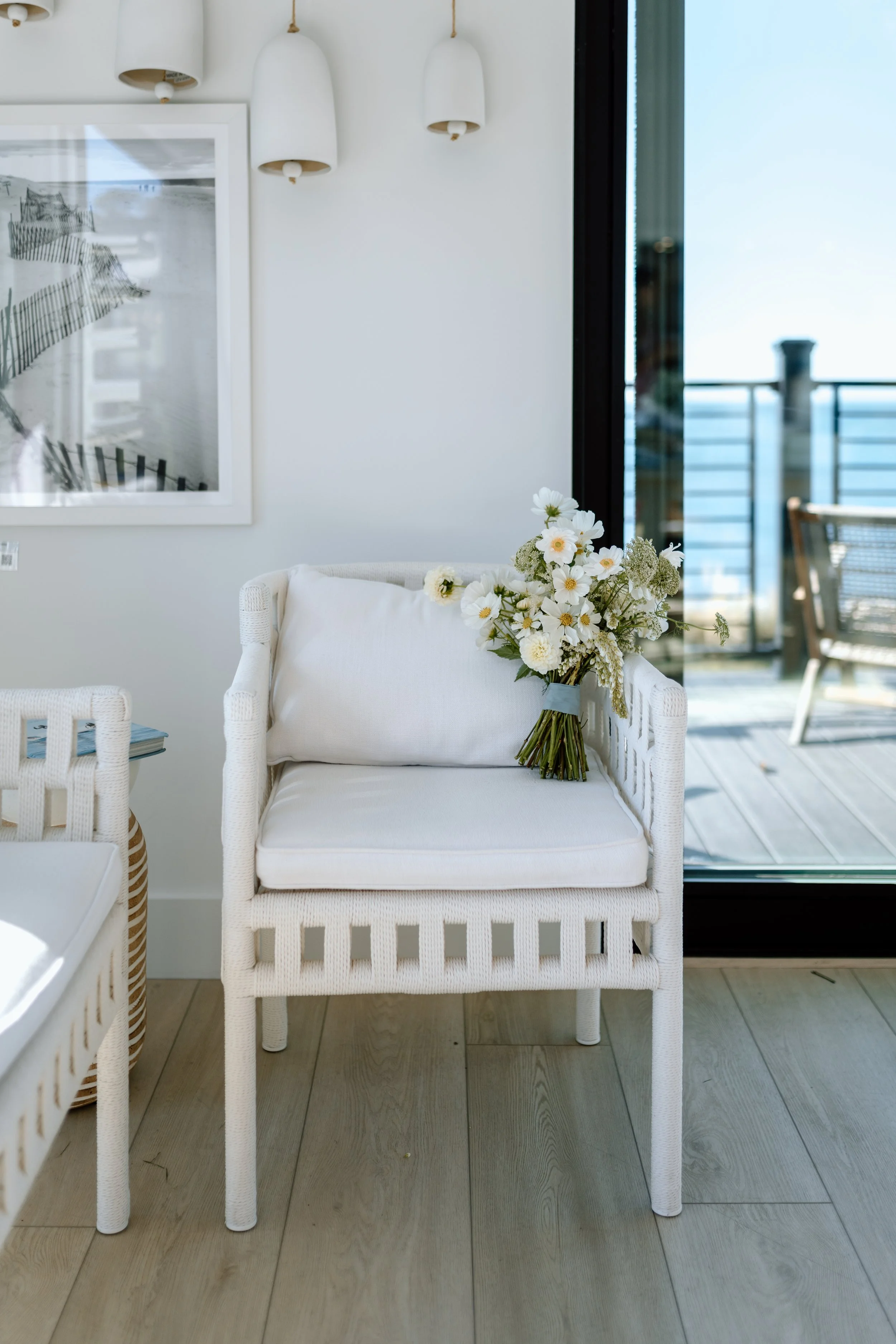 A white wicker chair with a cushion and pillow, holding a bouquet of white flowers, in a bright indoor space near a sliding glass door leading to a balcony with outdoor furniture and ocean view.