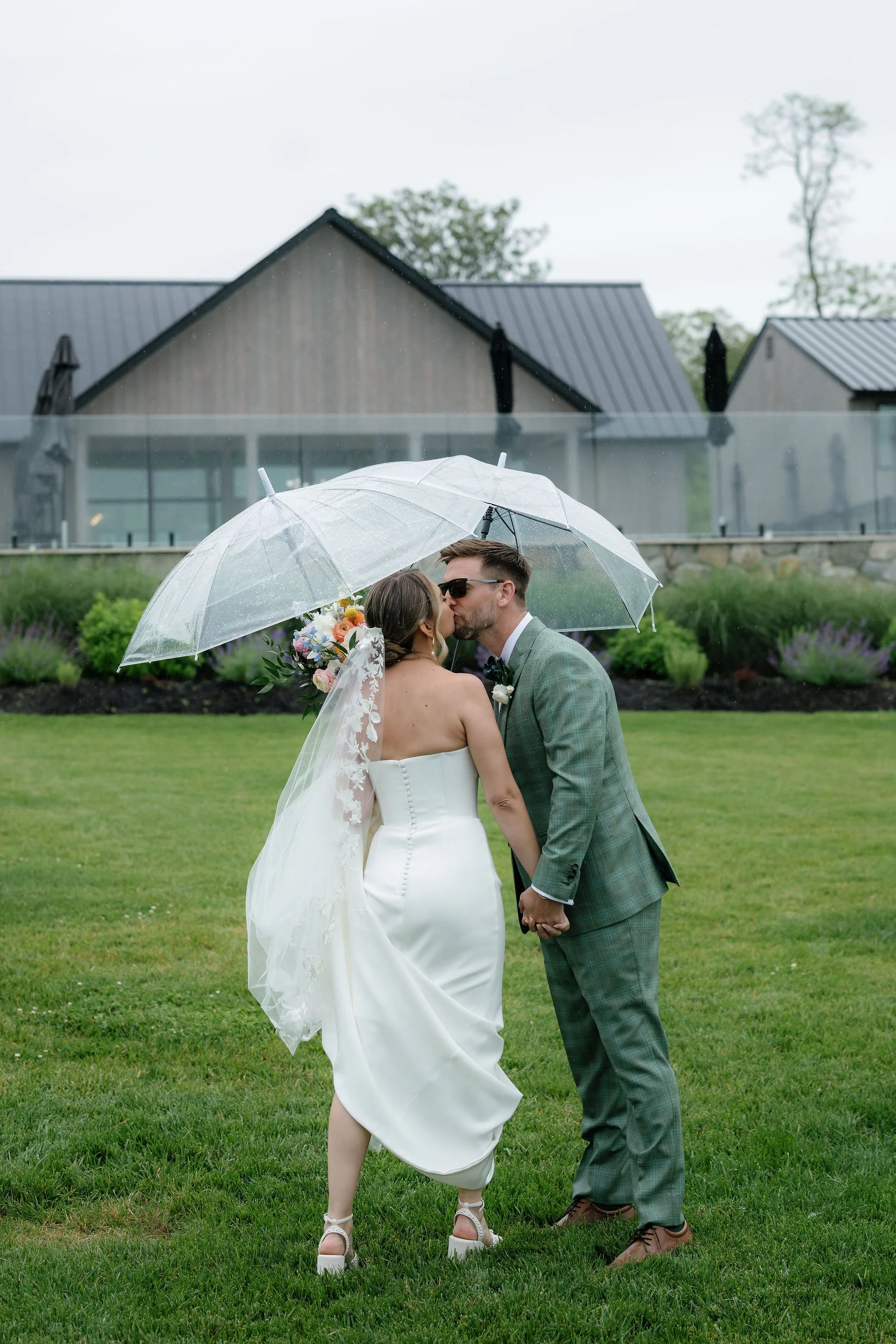 A bride and groom kiss under a clear umbrella on a grassy lawn, with rustic modern buildings and bushes in the background.
