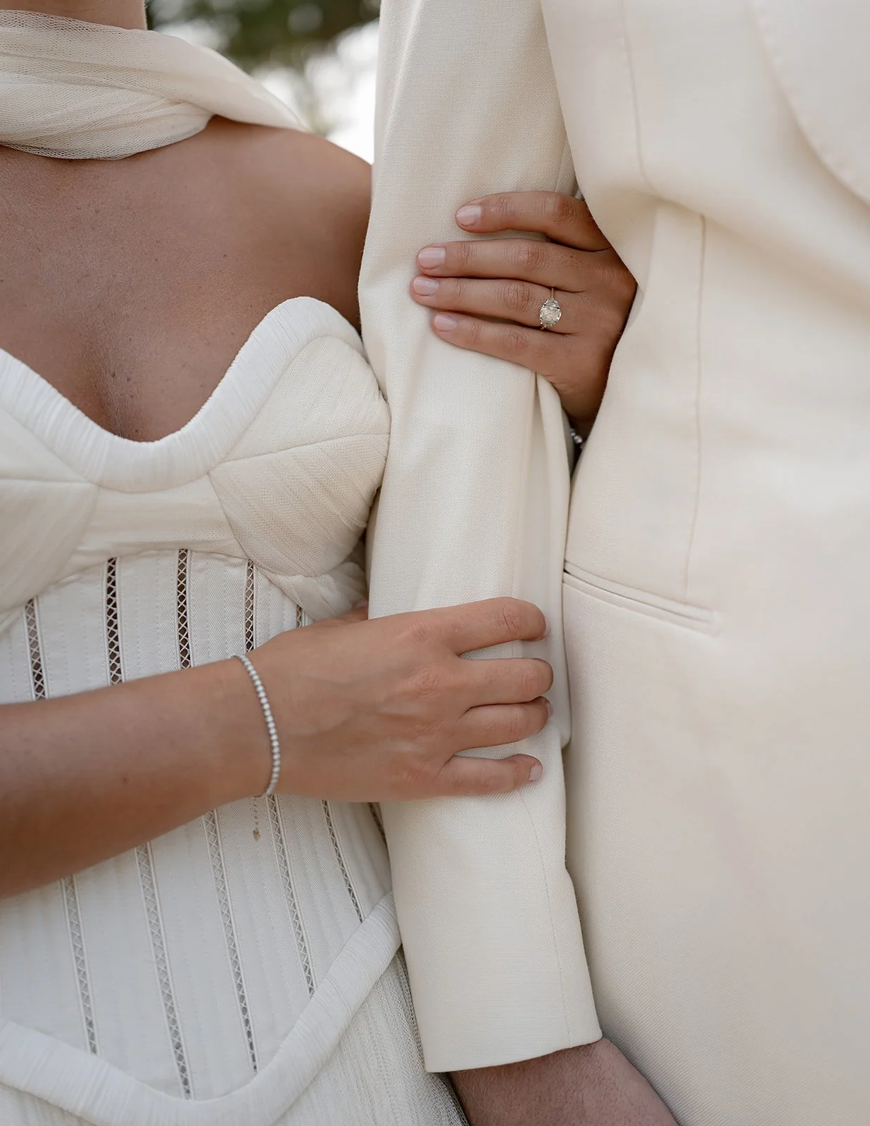Close-up of a couple's hands and arms, one woman with an engagement ring, embracing, wearing white clothing, against a blurred outdoor background.
