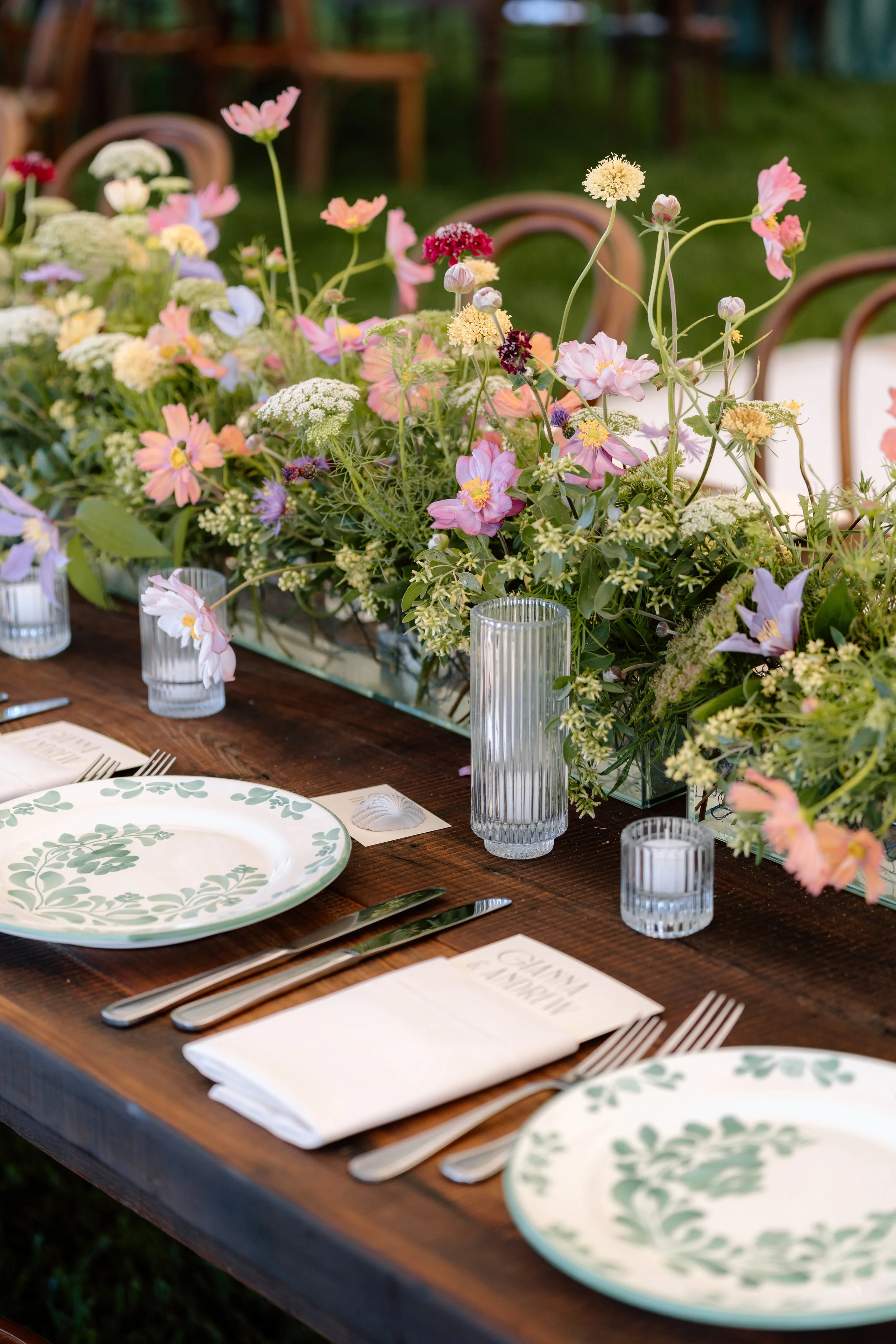 Table setup with floral arrangement, plates, napkins, and glassware at an outdoor event.