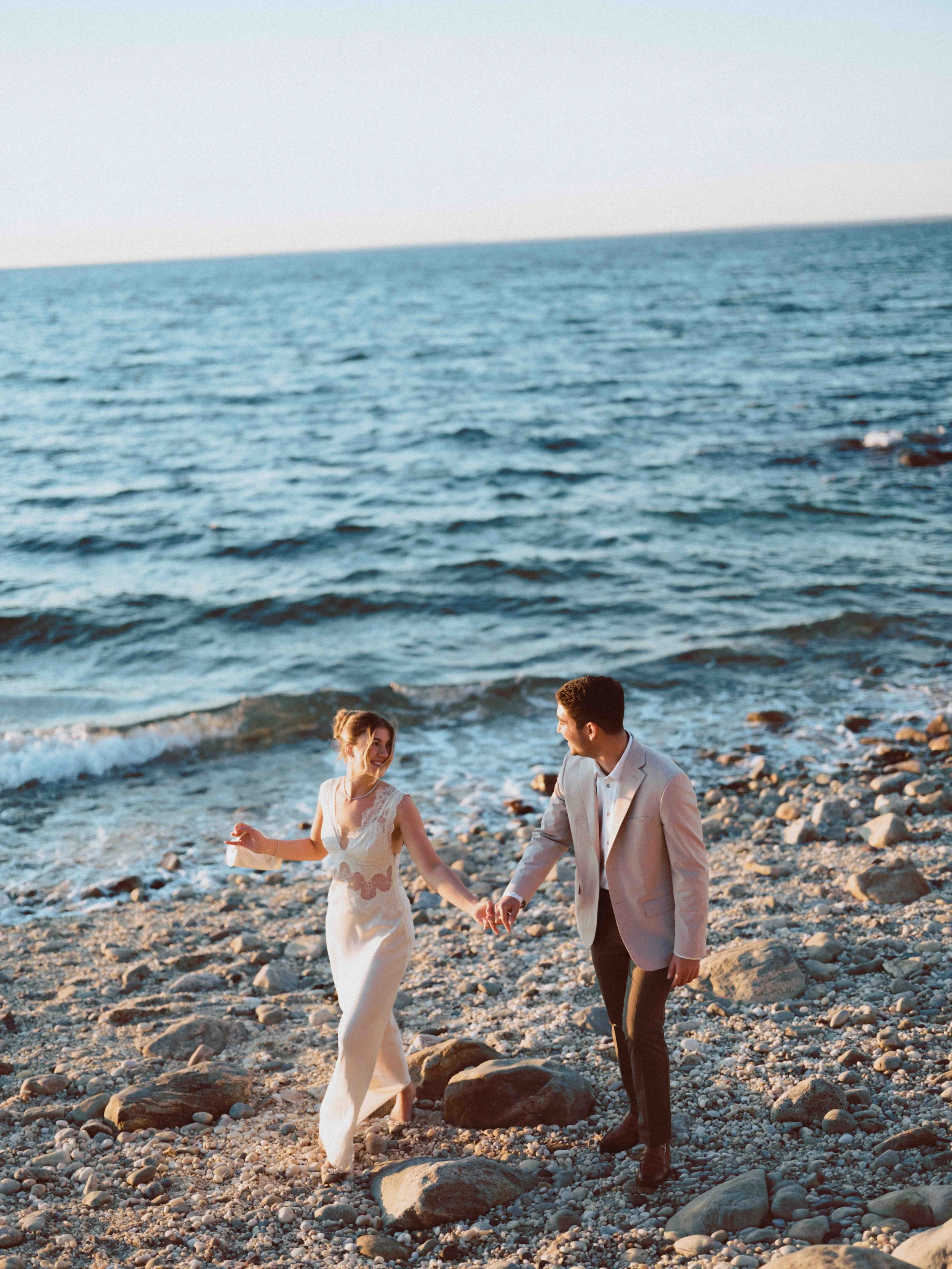 A couple dressed in wedding attire walking hand in hand on a rocky beach at sunset with the ocean in the background.