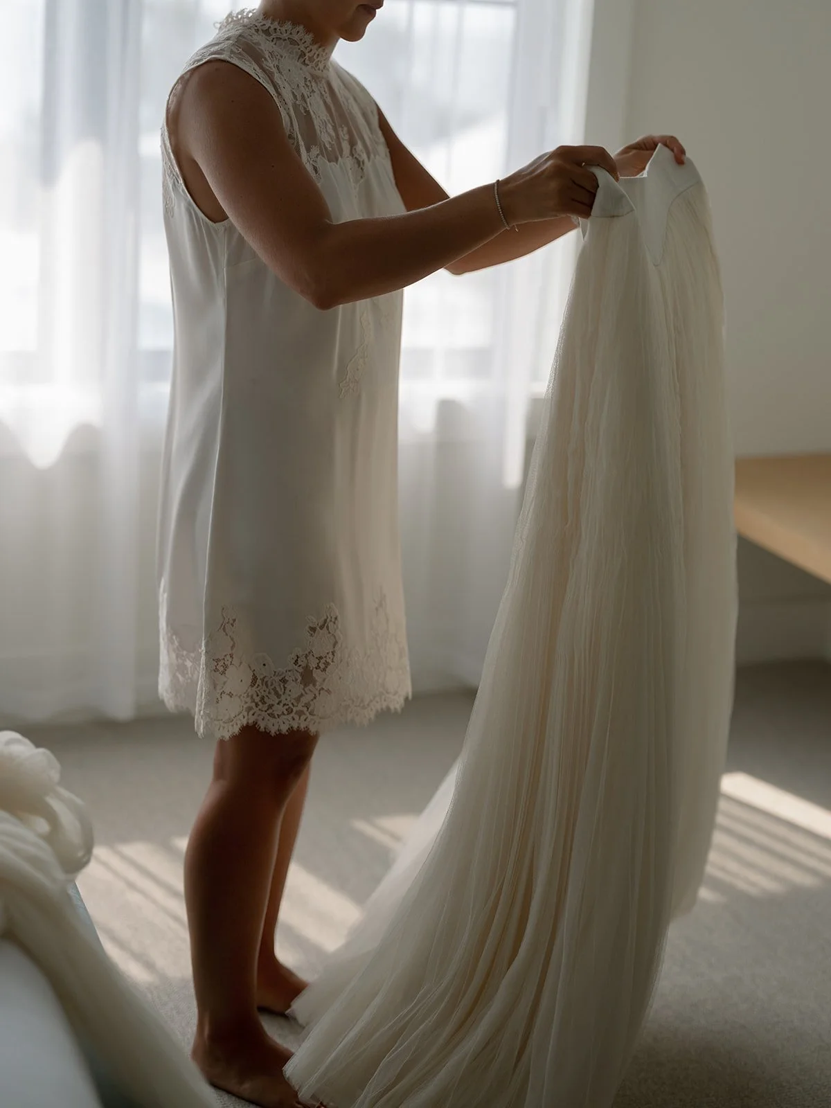 A woman in a white lace dress holding a cream-colored wedding gown indoors with natural light coming through sheer curtains.