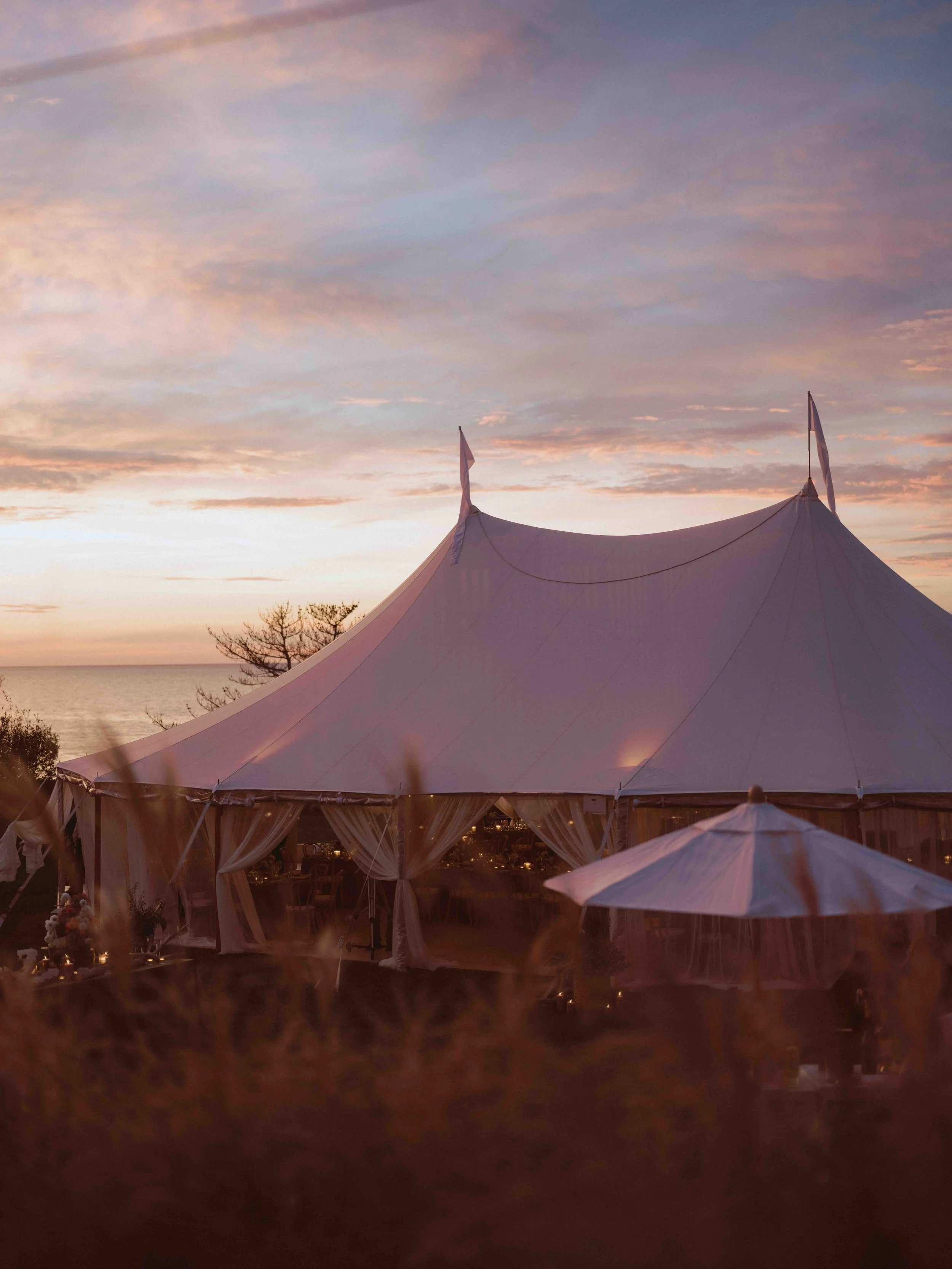 A large white event tent with draped curtains set up outdoors during sunset, with an ocean in the background and a sky filled with soft pink and purple clouds.