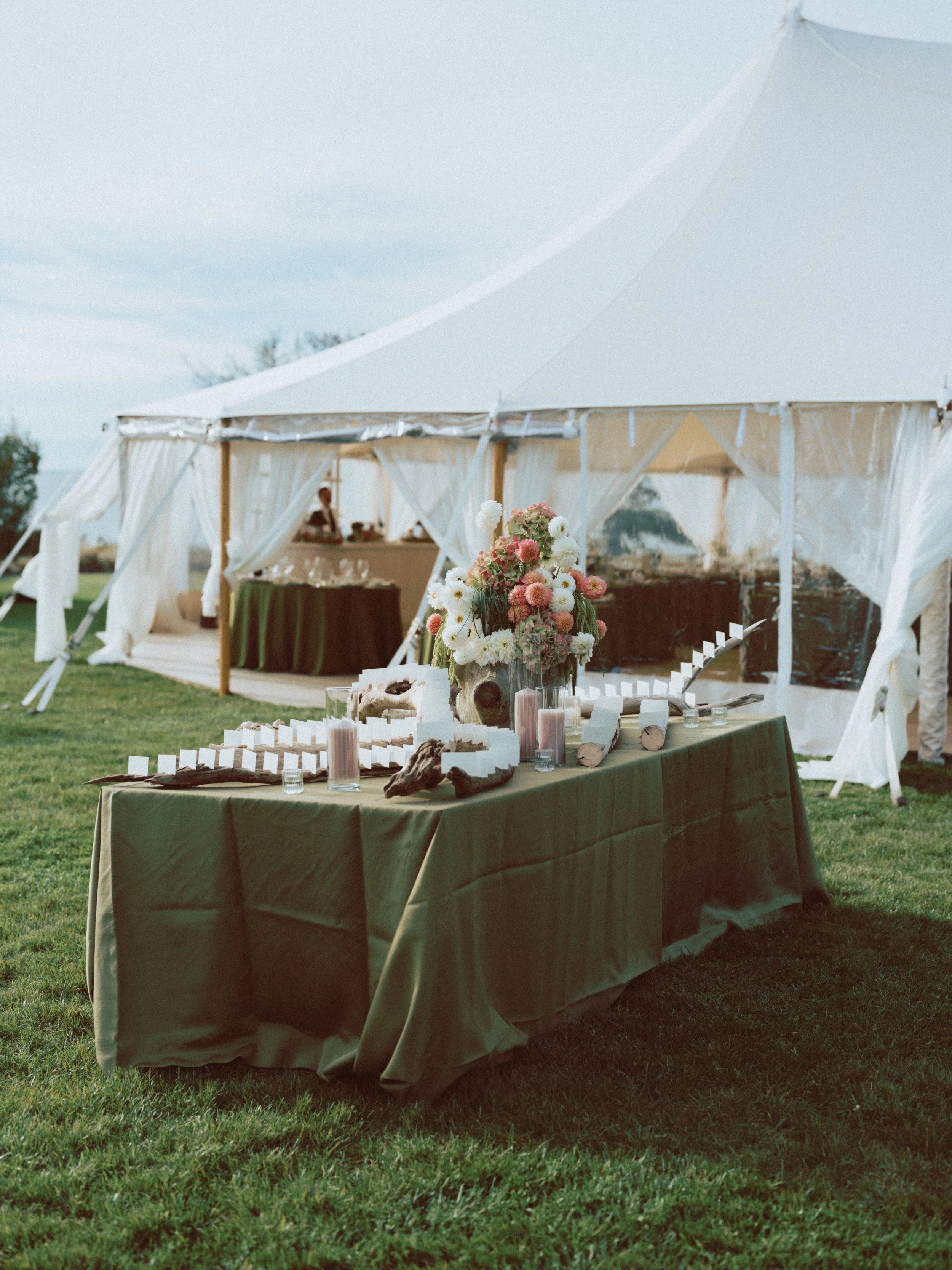 Decorative outdoor event setup with a table covered in a green cloth, centerpiece of pink and white flowers, candles, logs, and small cards, with a white event tent in the background.