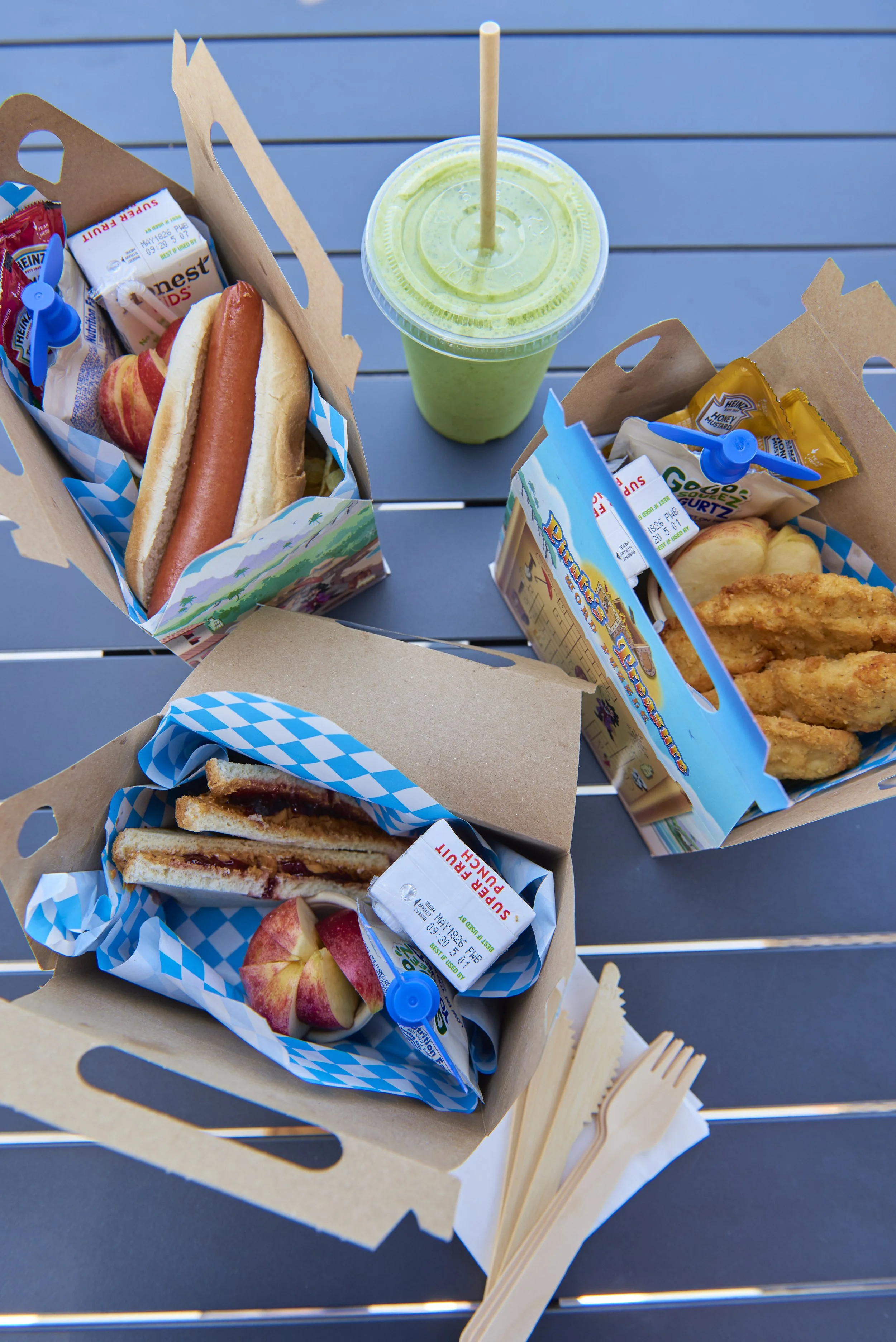 A spread of food and drinks on a black table including 3 Hotel Moraine signature "Matey Meals" including a Hot Dog, Chicken Fingers, Peanut Butter + Jelly Sandwich along with a green smoothie