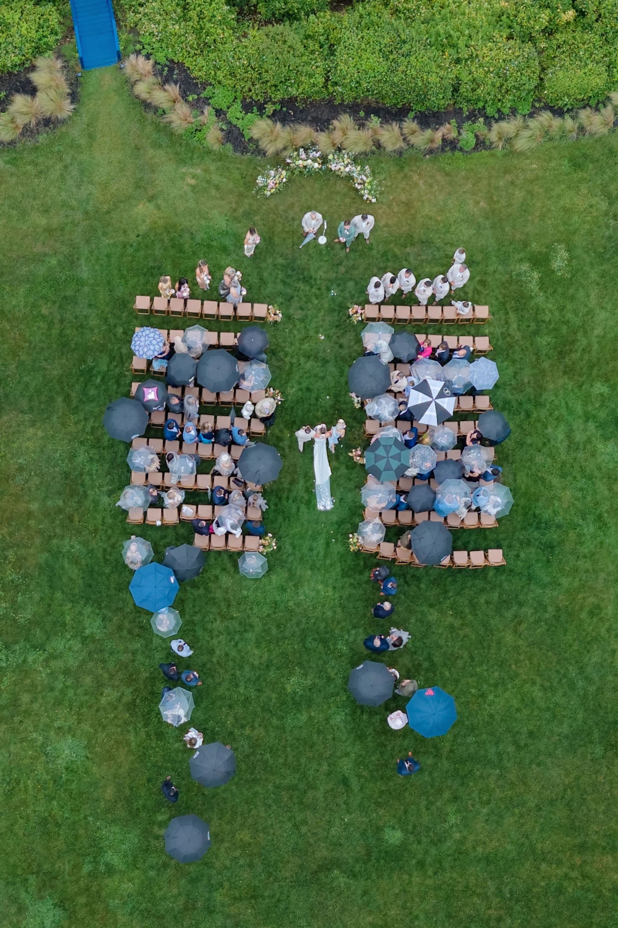 An outdoor wedding ceremony setup on a grassy lawn with rows of chairs and umbrellas, a bride and groom at the altar, guests seated, and a group of people standing at the front.