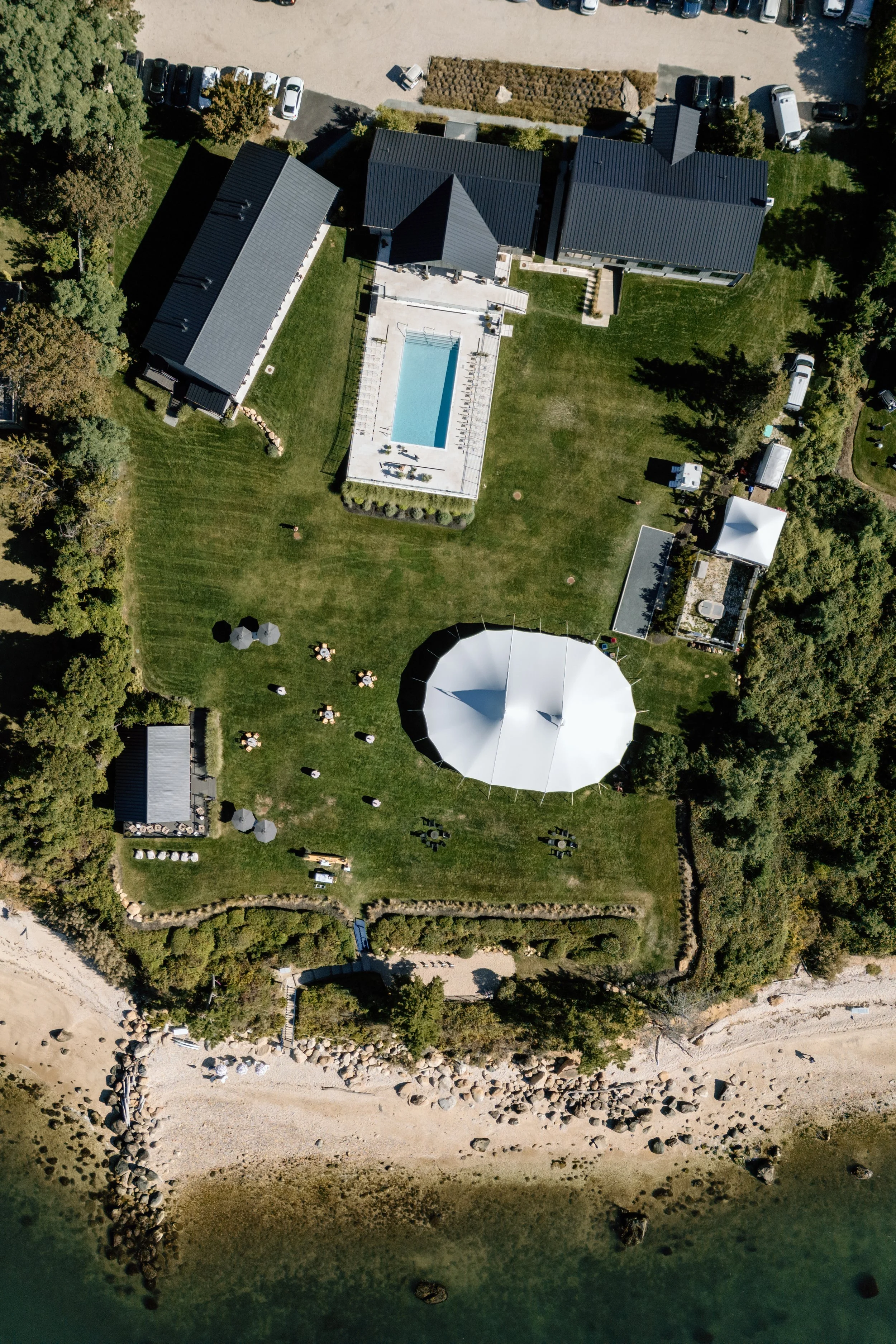 Aerial view of a large outdoor area featuring a swimming pool, a round pavilion, several tents, and multiple small tables with umbrellas on a grassy lawn, with surrounding trees, buildings, and a rocky shoreline along the water.