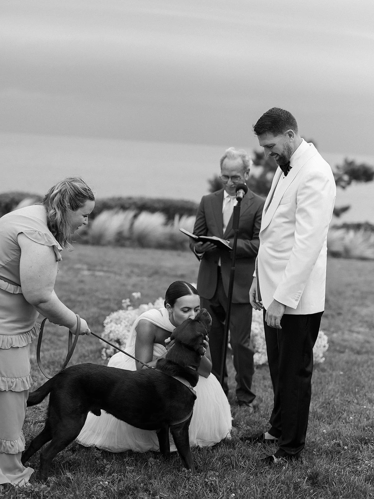 A wedding ceremony outdoors with a bride, groom, officiant, and a woman with a dog on a leash. The bride is kissing the dog, and the groom is smiling.