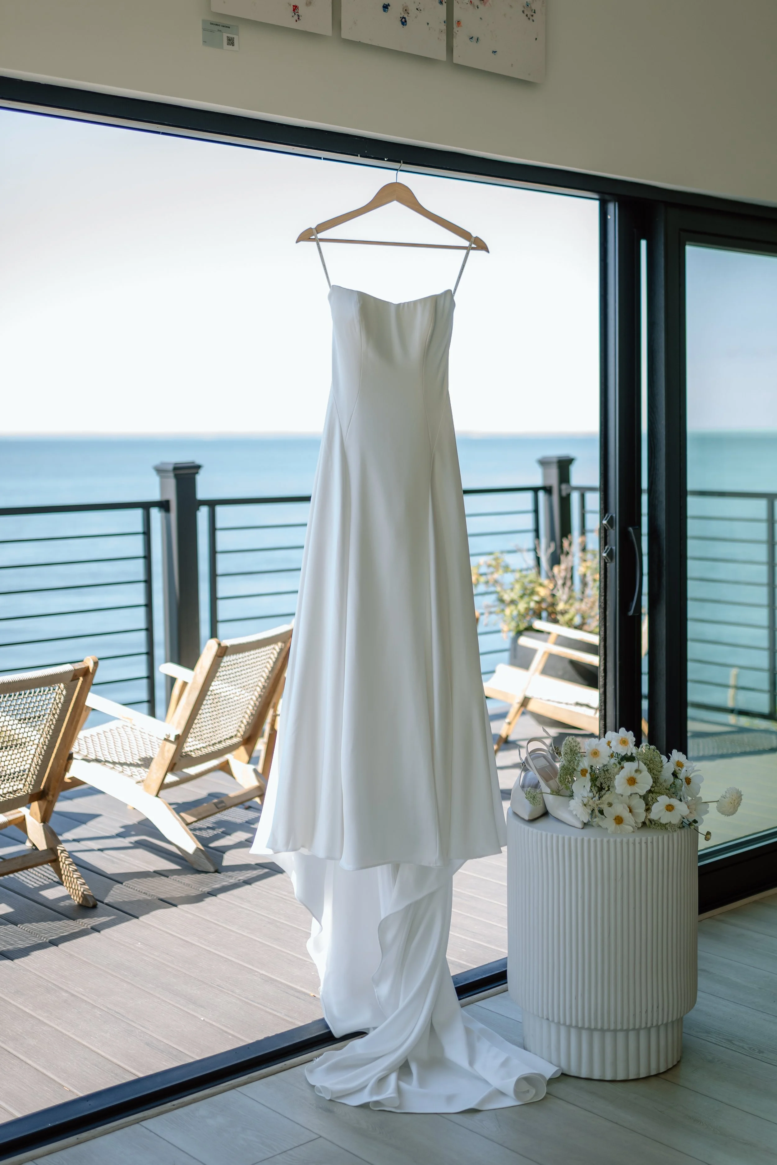 White wedding dress hanging on a wooden hanger in front of a sliding glass door, with a view of the ocean and outdoor chairs on a balcony, and a bouquet of white flowers on a white ribbed pedestal.