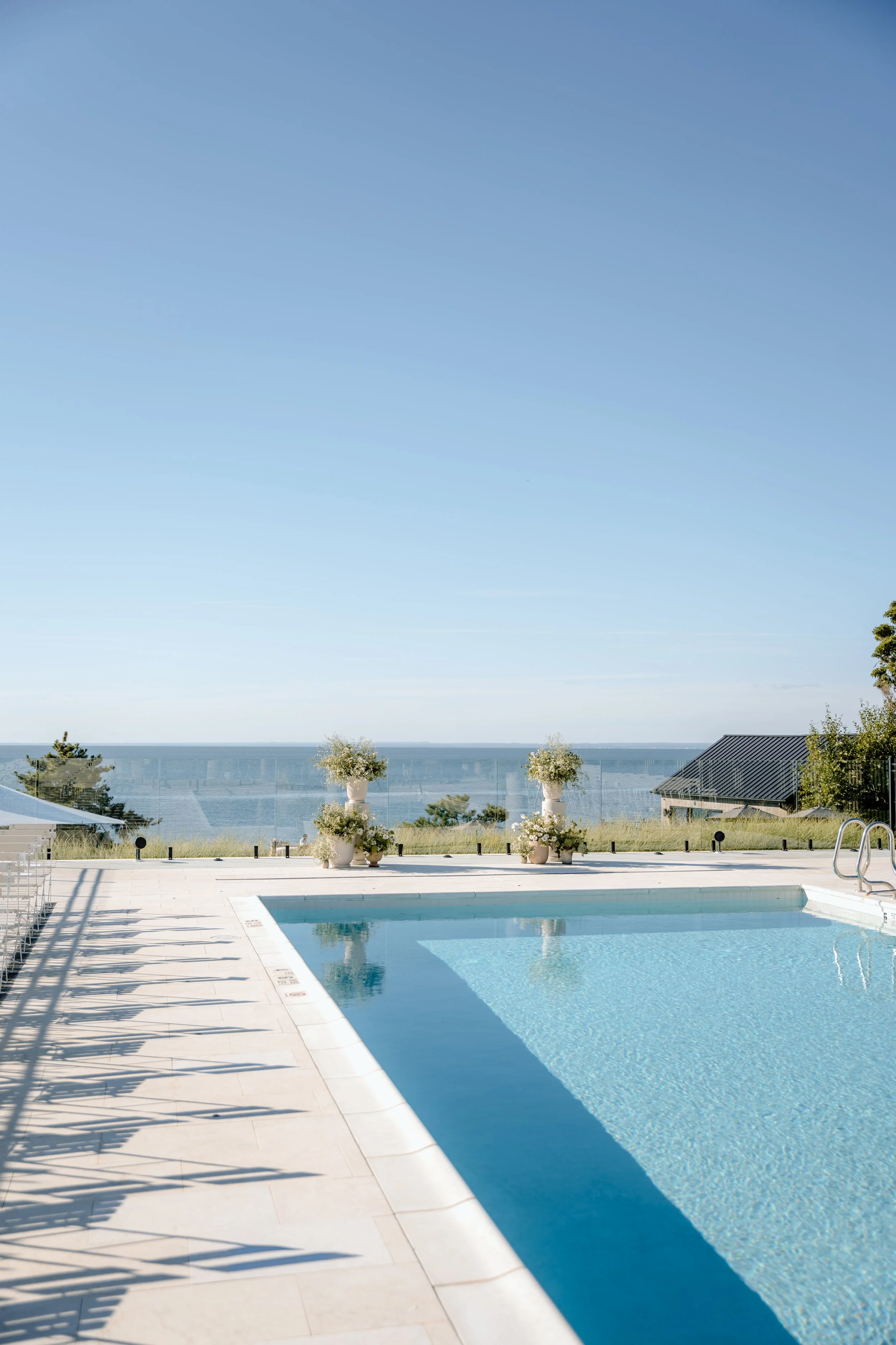 Outdoor swimming pool with a view of the ocean, potted plants with white flowers, clear blue sky.