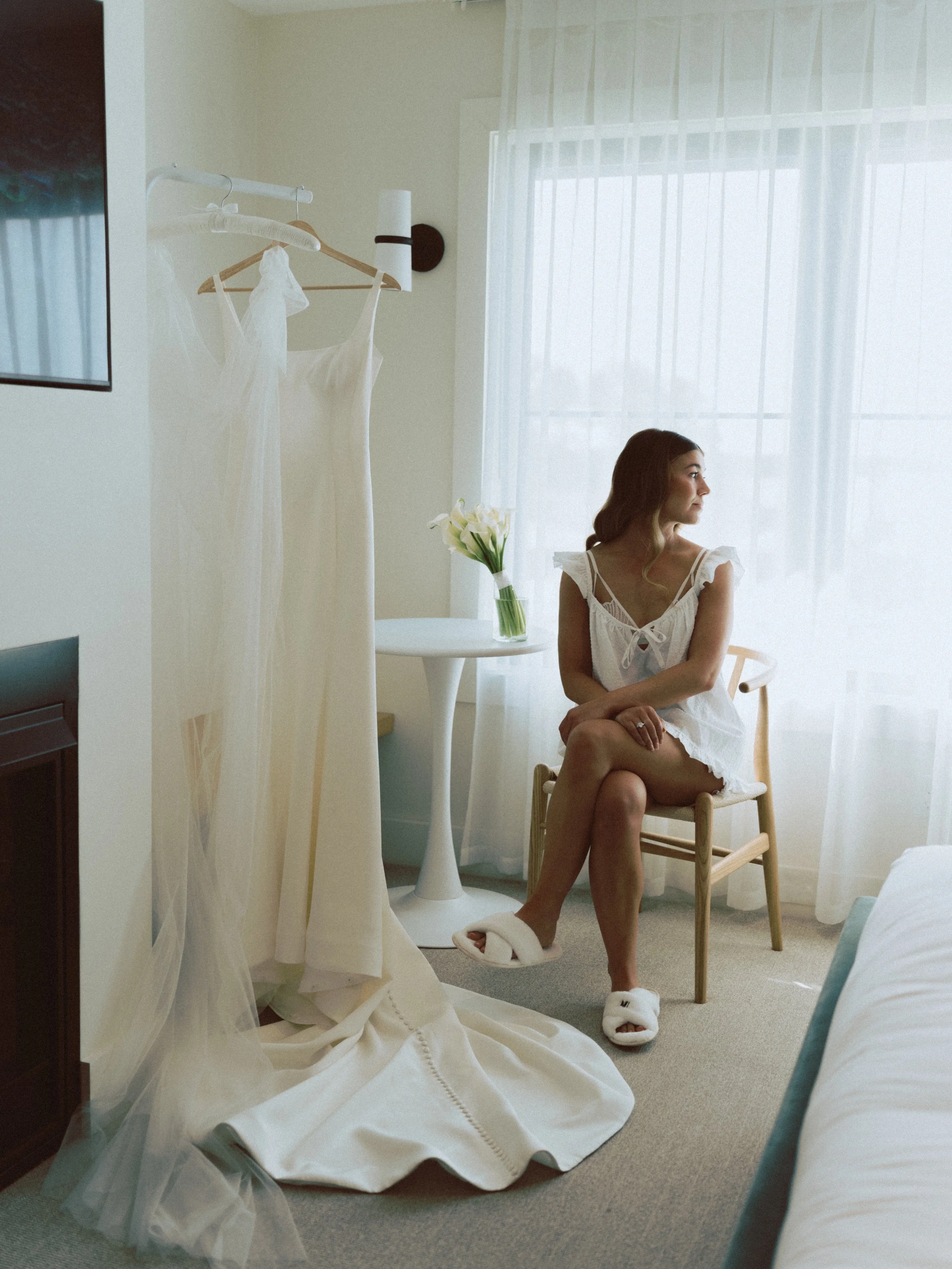 A woman sitting on a chair in a hotel room, looking outside the window. Two wedding dresses hang on a rack nearby, and a bouquet of white flowers sits on a table