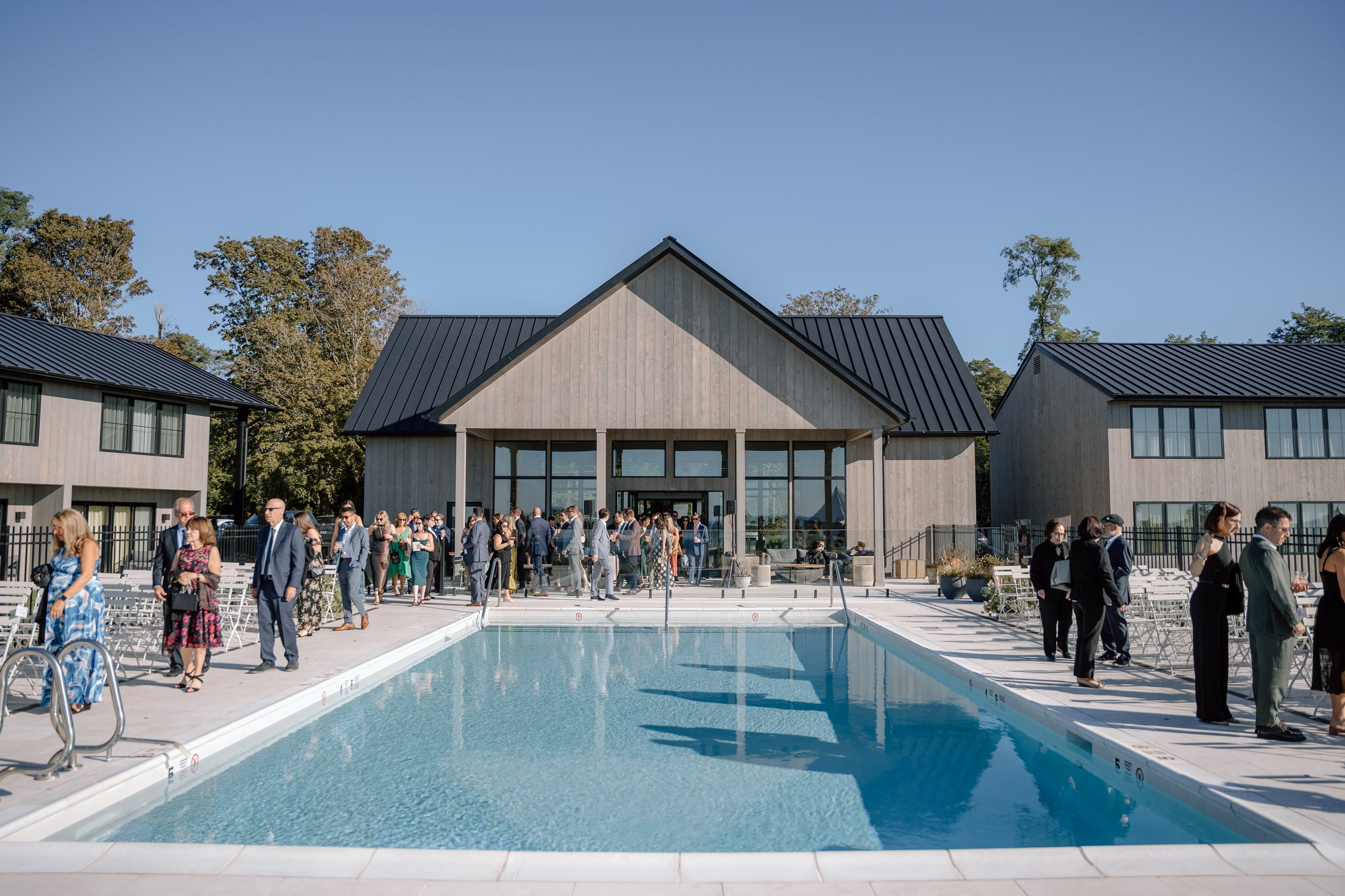 People dressed in formal attire gather around a swimming pool outside a modern building with wooden siding and a black metal roof during a sunny day.