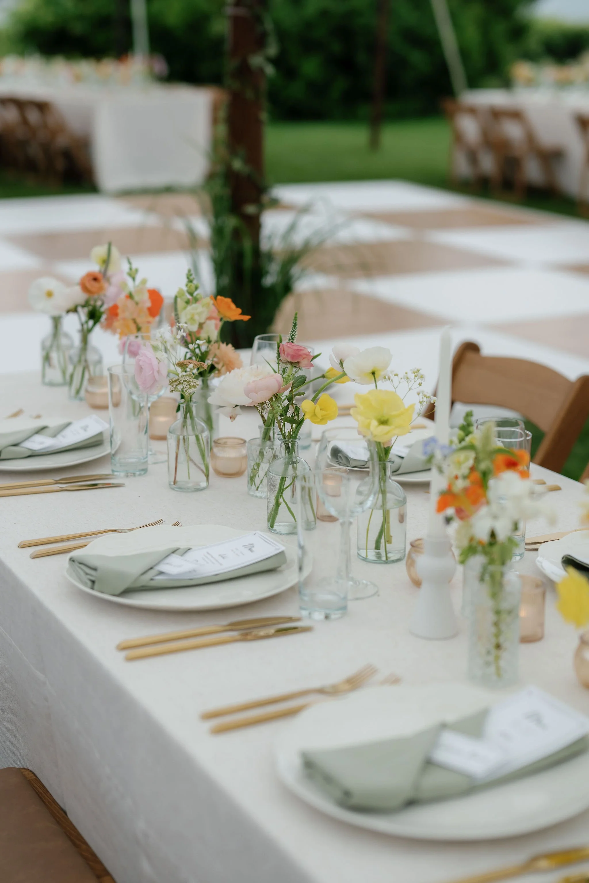 Table set for a wedding or event outdoors, decorated with a white tablecloth, floral centerpieces in glass vases, candles, plates, napkins, and gold flatware, with a grassy area and trees in the background.