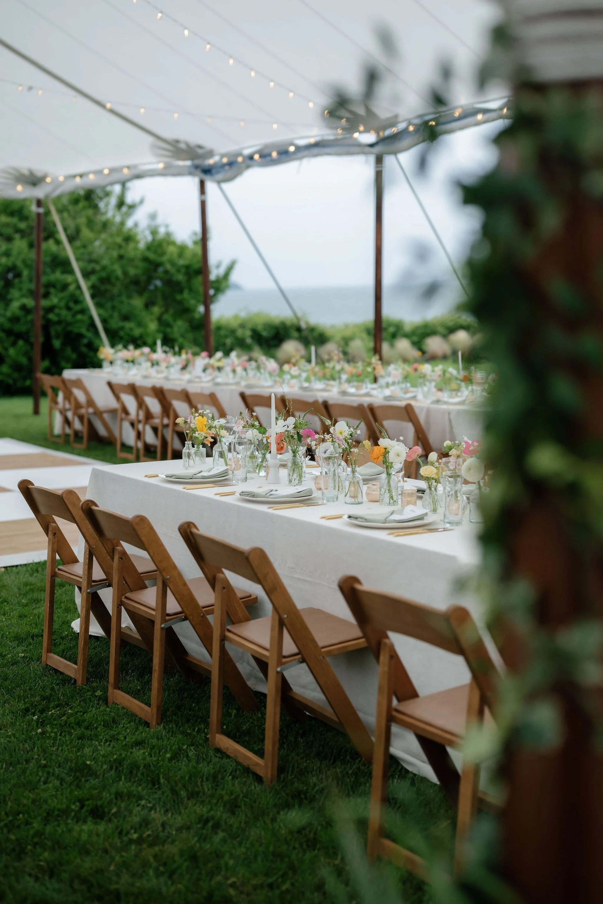 Decorated outdoor dining setup with long tables, floral arrangements, and string lights under a canopy, overlooking a green lawn and water in the distance.