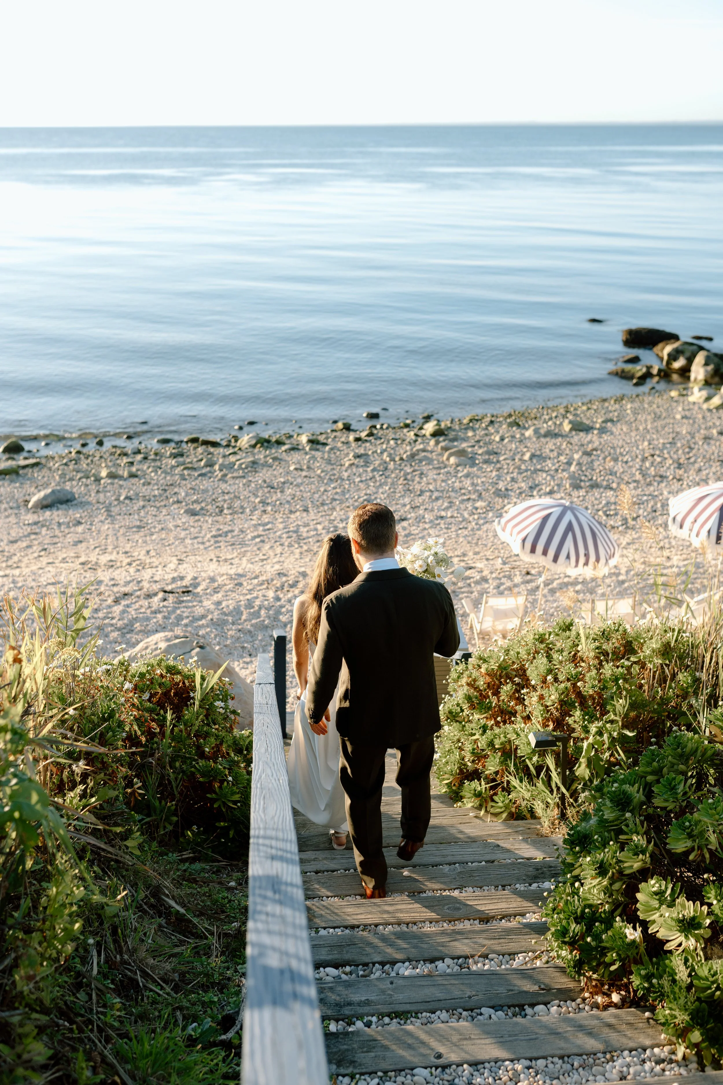 A couple dressed in wedding attire walking down a wooden staircase towards a beach, with umbrellas and lounge chairs on the sand, leading to a calm ocean.