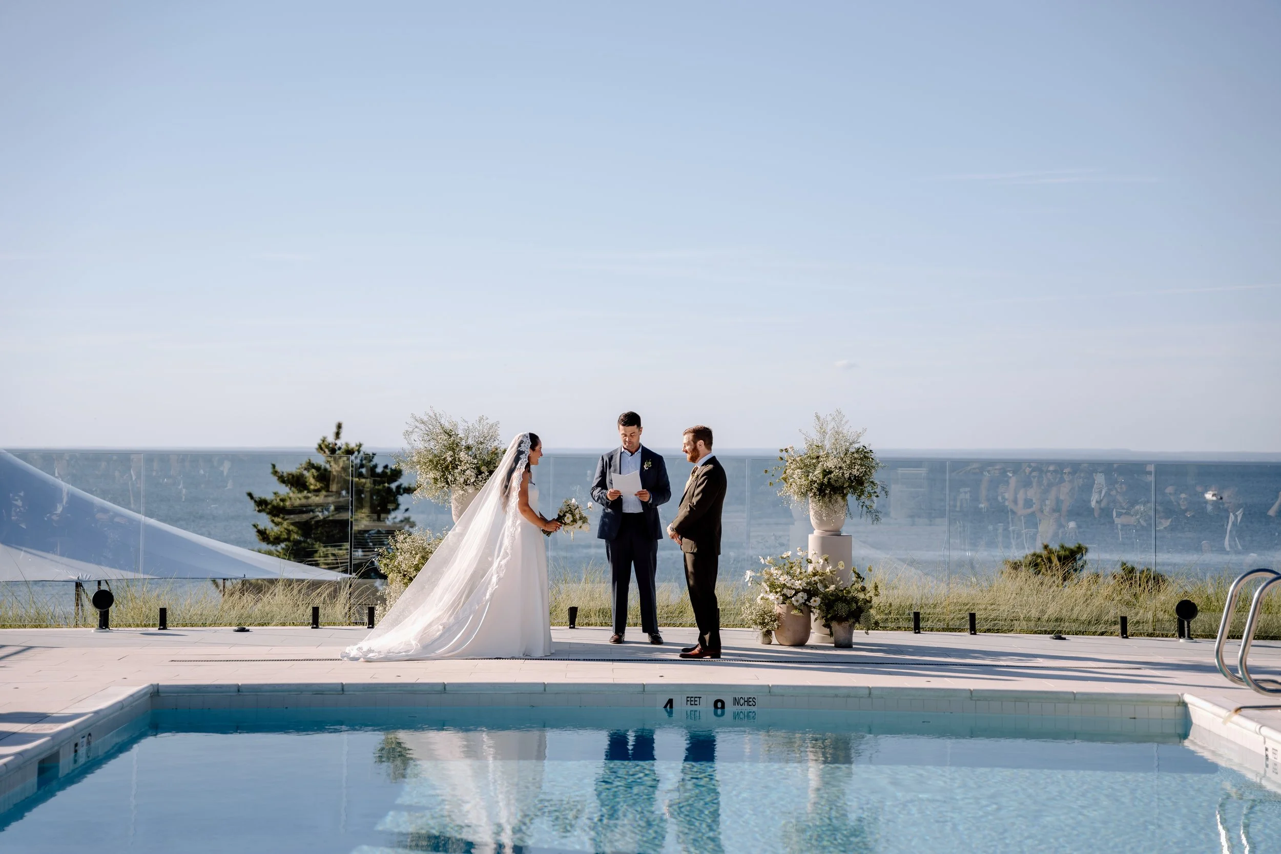 A bride and groom exchanging vows during a wedding ceremony by a pool with a view of the ocean, decorated with floral arrangements.