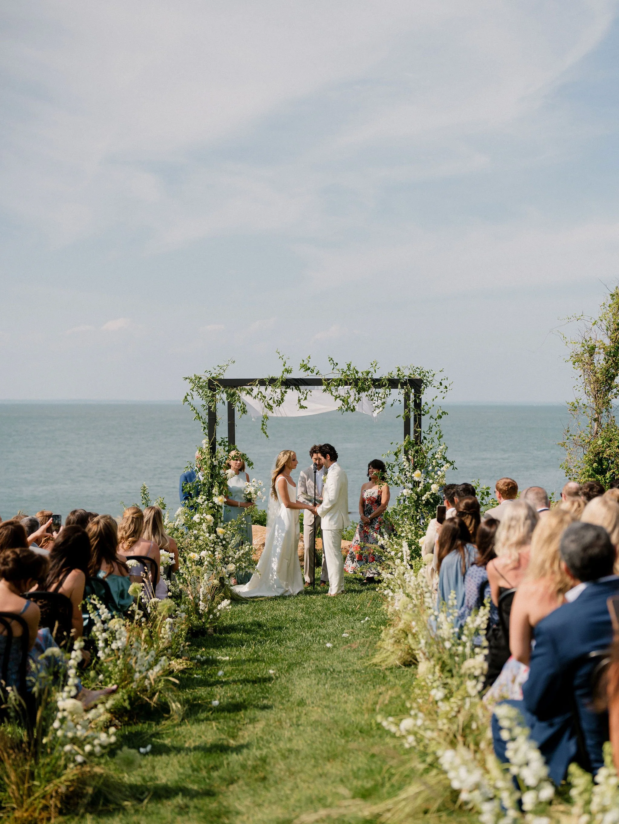 A wedding ceremony taking place outdoors by the ocean with a bride and groom holding hands at the altar, surrounded by flower arrangements and seated guests.