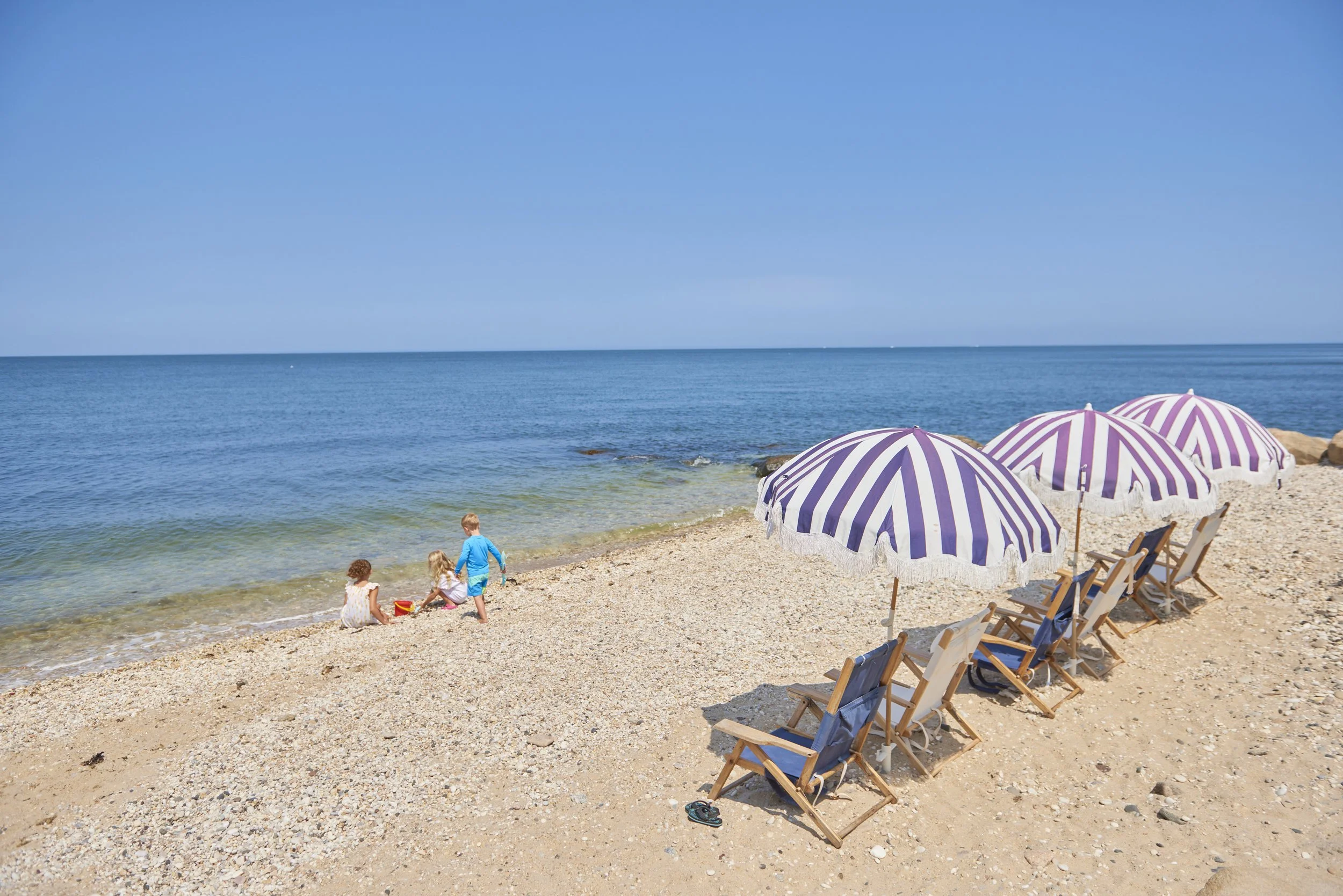 A scene of children playing at the waters edge on the beach at Hotel Moraine with the Long Island Sounds in the distance. Lounge chairs and striped beach umbrellas are set up on the rocky-sandy beach
