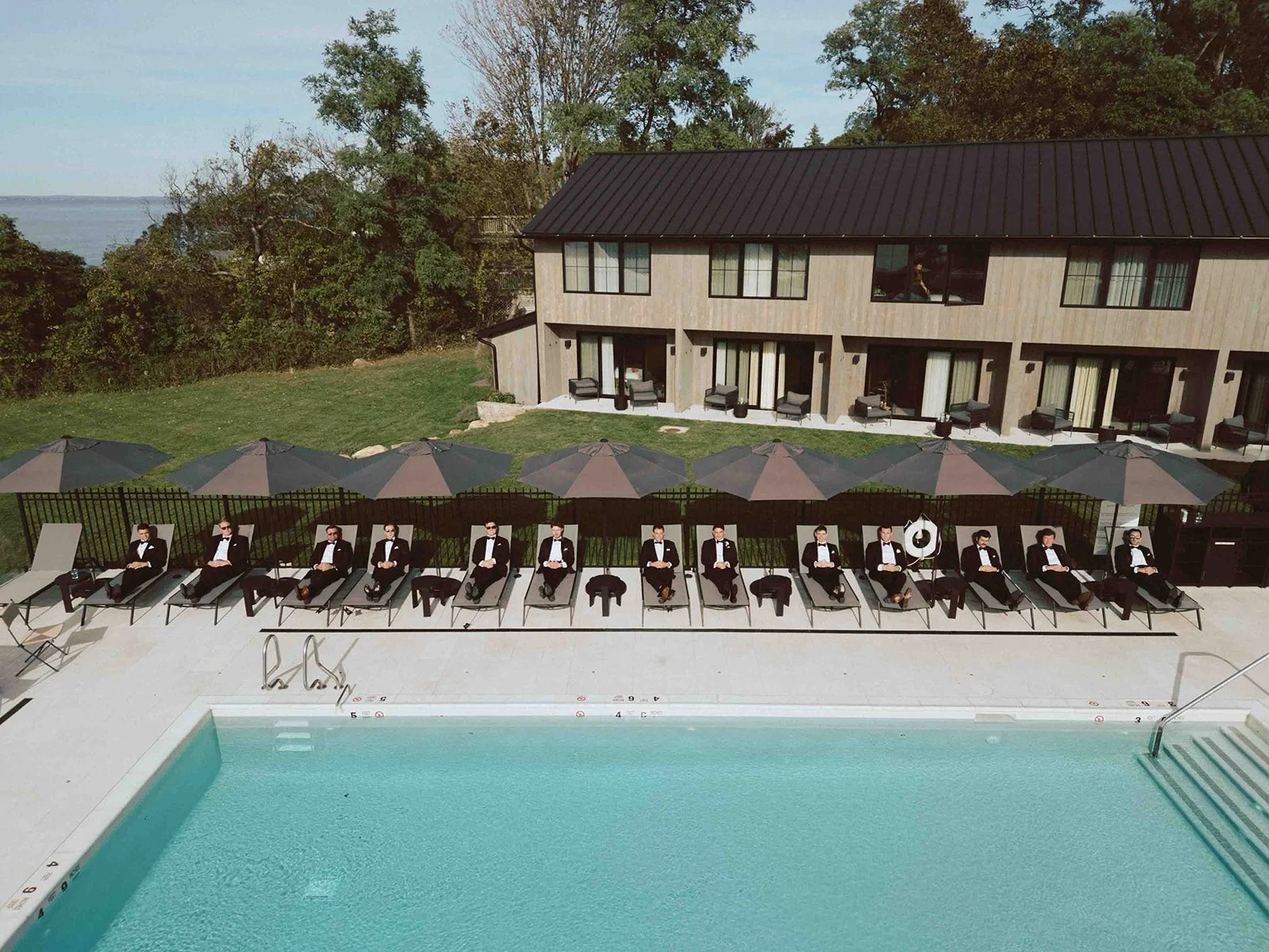 Ten men in tuxedos and sunglasses relaxing on lounge chairs beside a swimming pool, with umbrellas overhead and a building with outdoor seating in the background.