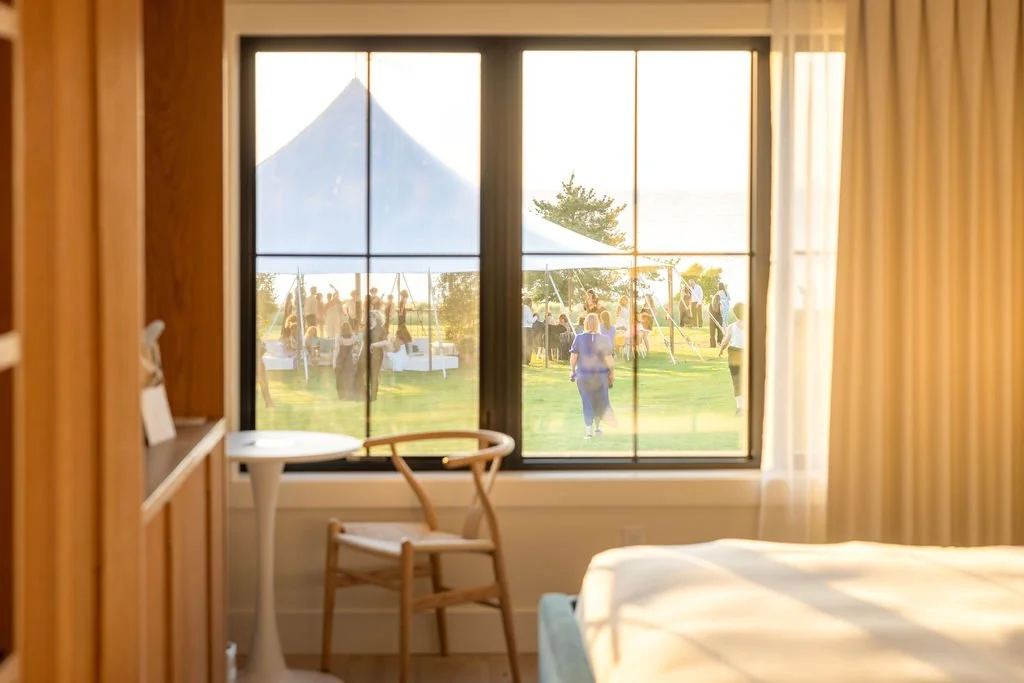 View from a bedroom window showing a gathering of people outside under a large tent on a grassy area, with mountains and trees in the background, during sunset.