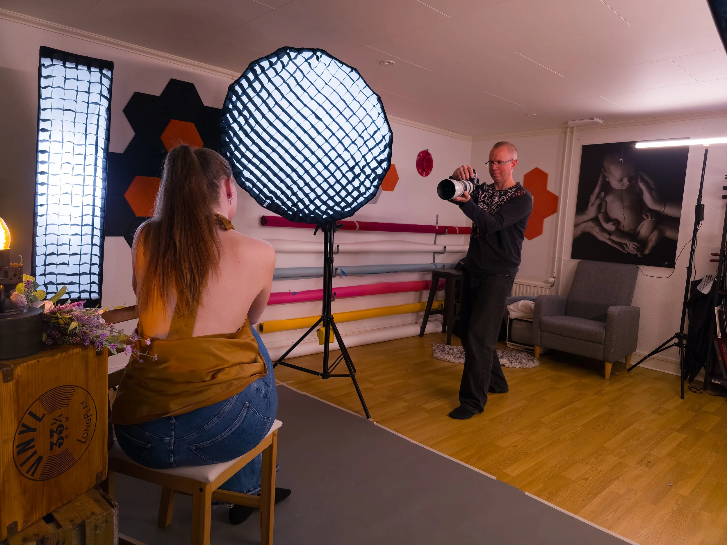 En fotograf som tar bilder av en modell i ett inomhusstudio med professionell belysning och bakgrund. Modellen sitter på en stol och har en gul jacka.