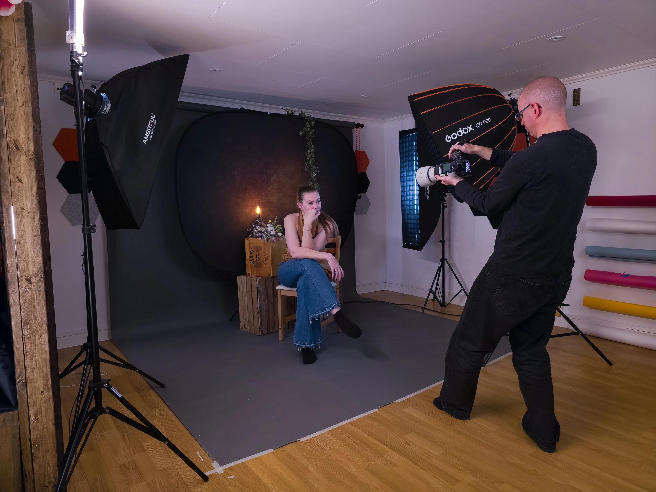 En fotograf tar porträtt av en ung kvinna som sitter på en stol med blomsterdekoration framför en svart bakgrund i ett fotostudio. Fotografen står bredvid och använder en stor blixt med paraply.