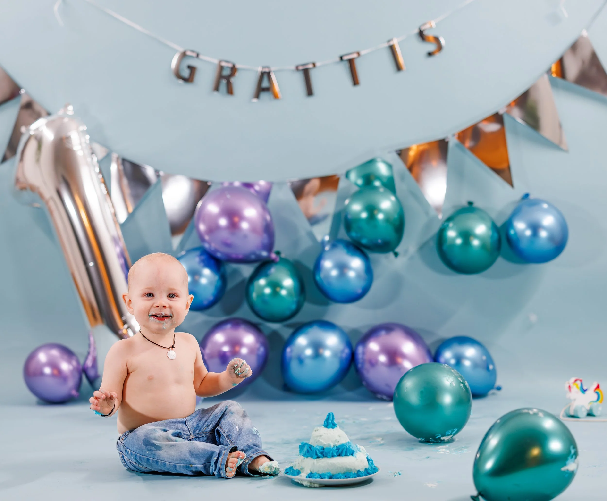 En pojke sitter på golvet med en tårta och äter. Bakom honom finns blå och lila ballonger, ett ”GRATTIS”-band och en födelsedagstema.