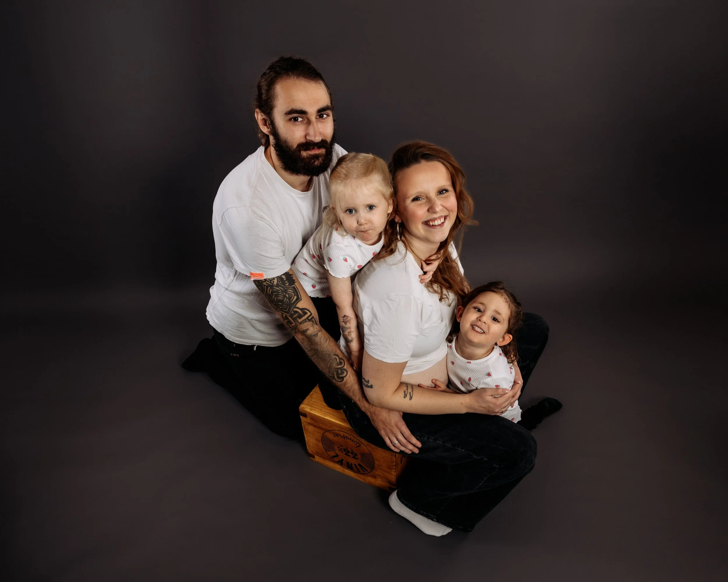 En familj med två vuxna och tre barn sitter på en träkloss mot en mörk bakgrund. Vuxna och barn ler och tittar mot kameran.