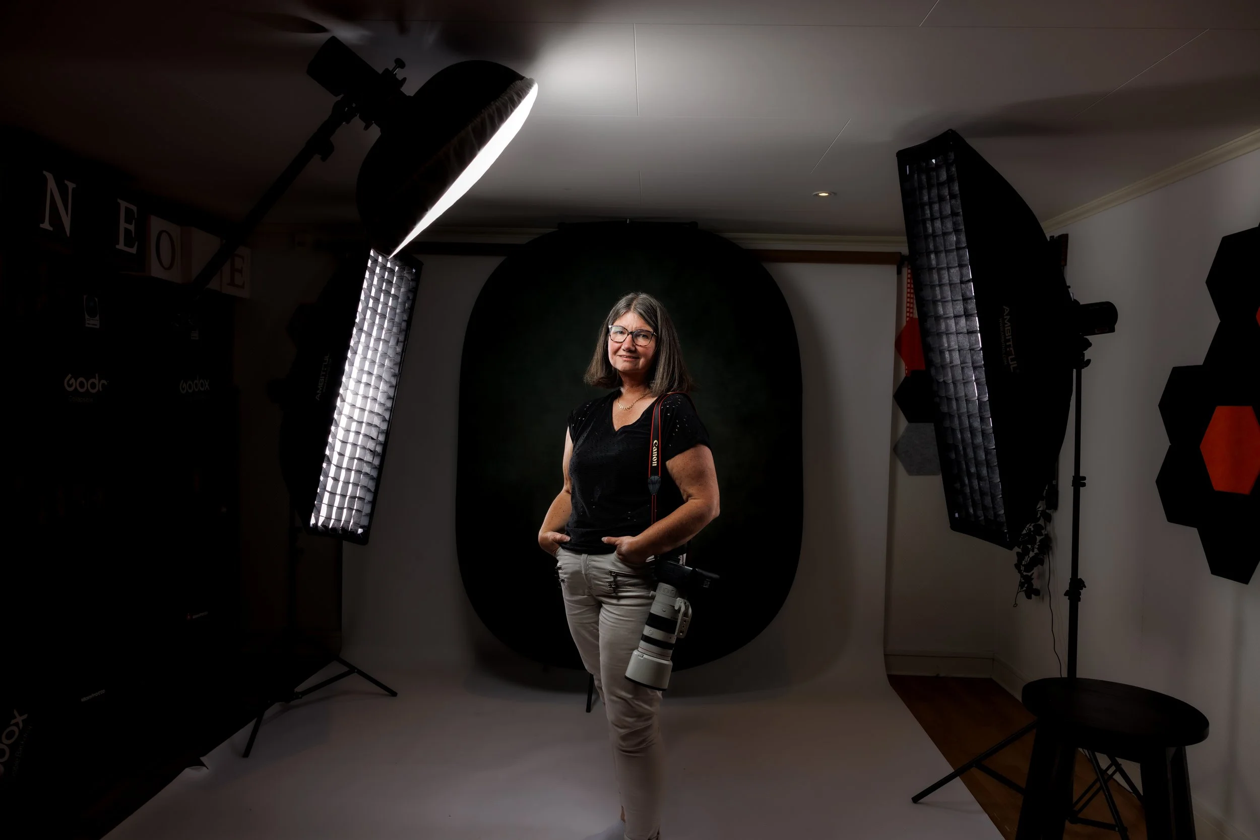 En kvinna med glasögon står i en fotostudio med fotografiutrustning, inklusive studiobelysning och ett enkelt svart bakgrundsark, och håller en stor teleobjektivkamera.