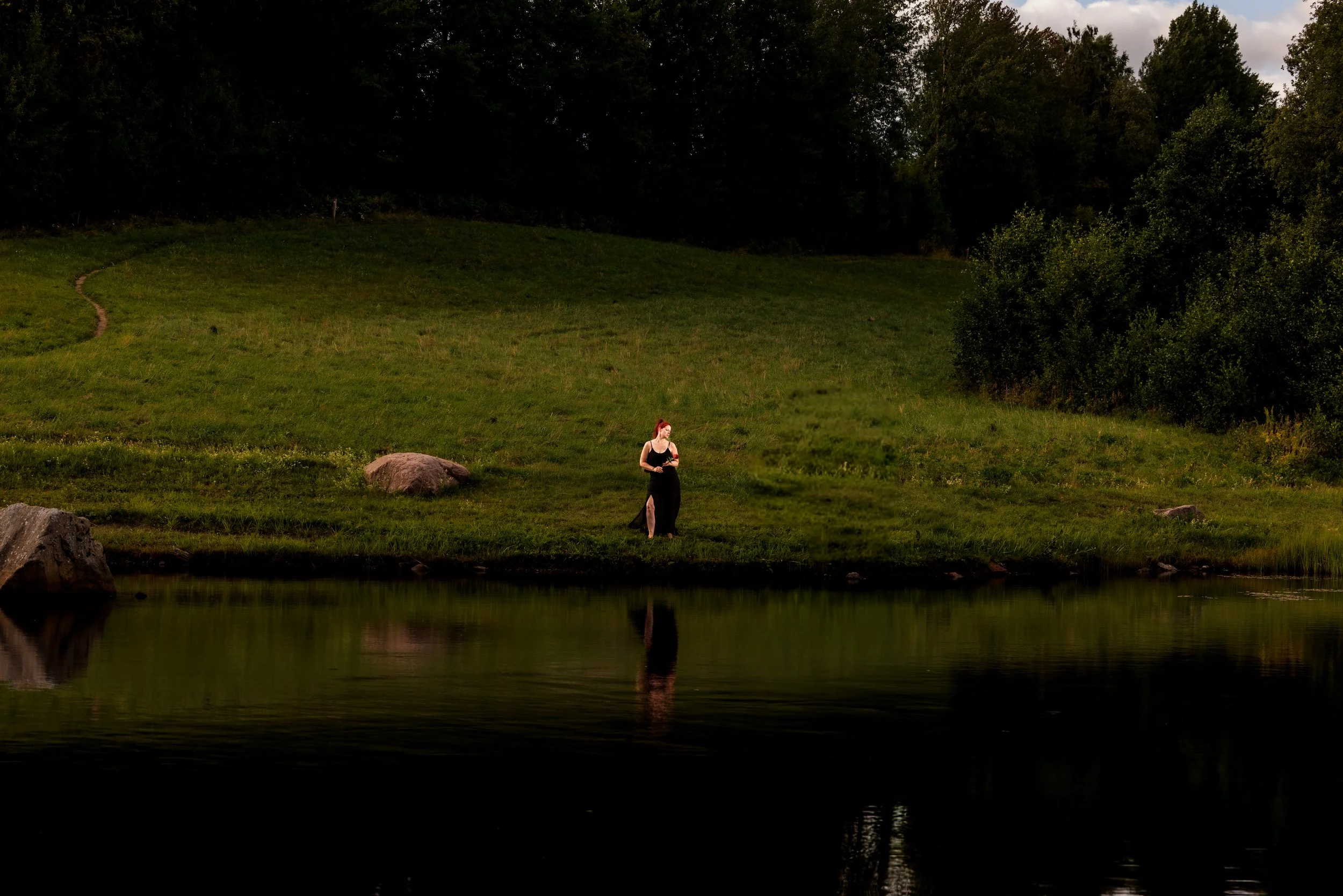 En kvinna i svart klänning står vid en flod omgiven av gröna träd, kvällssol eller skymning, reflekterad i vattnet.