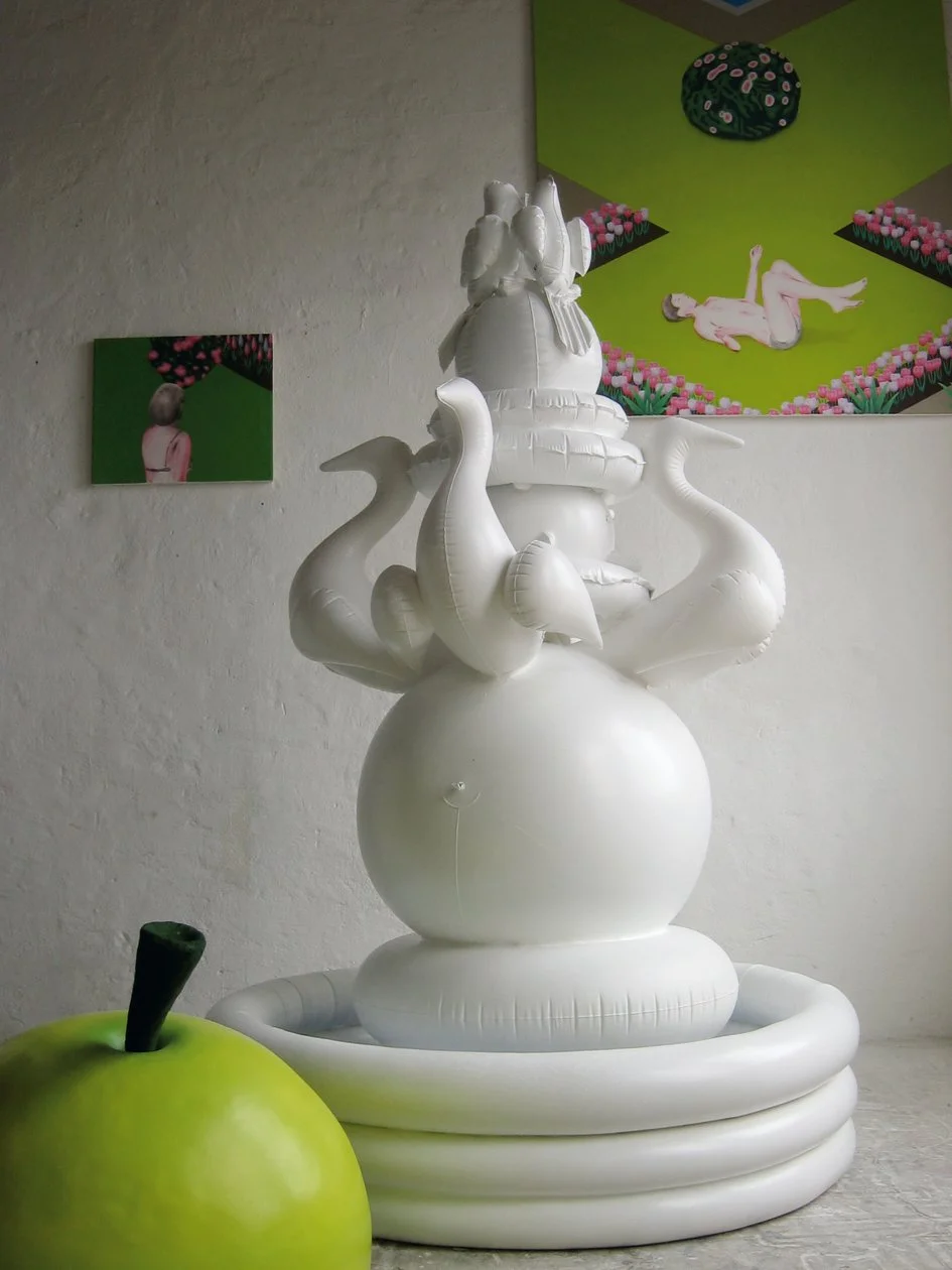 Ein angeschnittener Apfel im Vordergrund  dahinter ein aufblasbarer Brunnen und zwei Bilder im HInetrgund auf der ein Mann auf dem Rücken liegend und der Torso einer Frau von Hinten zu sehen ist